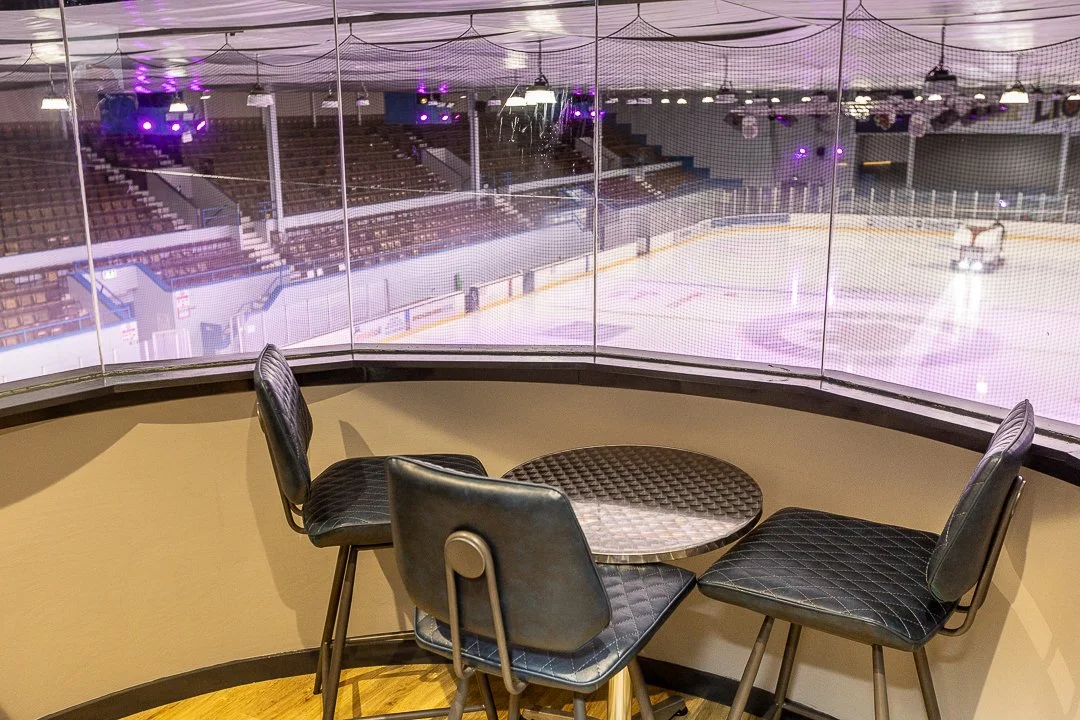 An indoor balcony overlooking an ice hockey rink with a small round table and four black chairs.