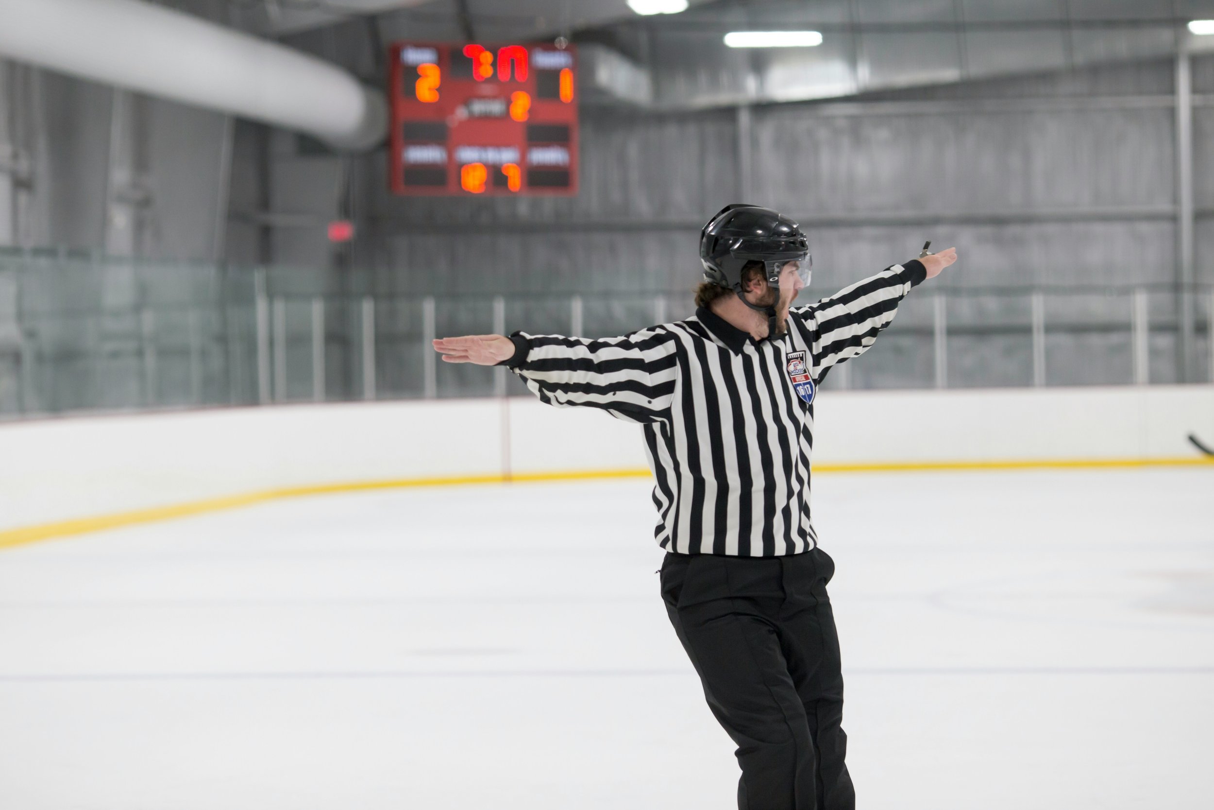 Hockey referee on the ice rink with arms outstretched, wearing a striped black and white uniform and black helmet, during a game with a digital scoreboard in the background.
