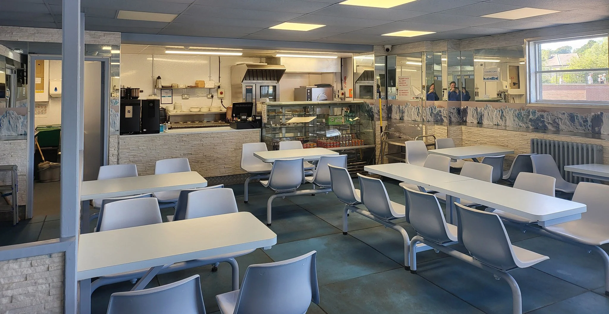 Empty cafeteria with white tables and chairs, serving counter in the background, and a large window to the right view outside.