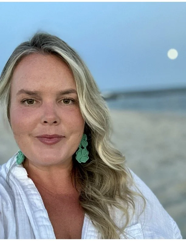 A woman with blonde hair wearing green earrings and a white shirt takes a selfie on the beach during sunset with the moon visible in the sky.