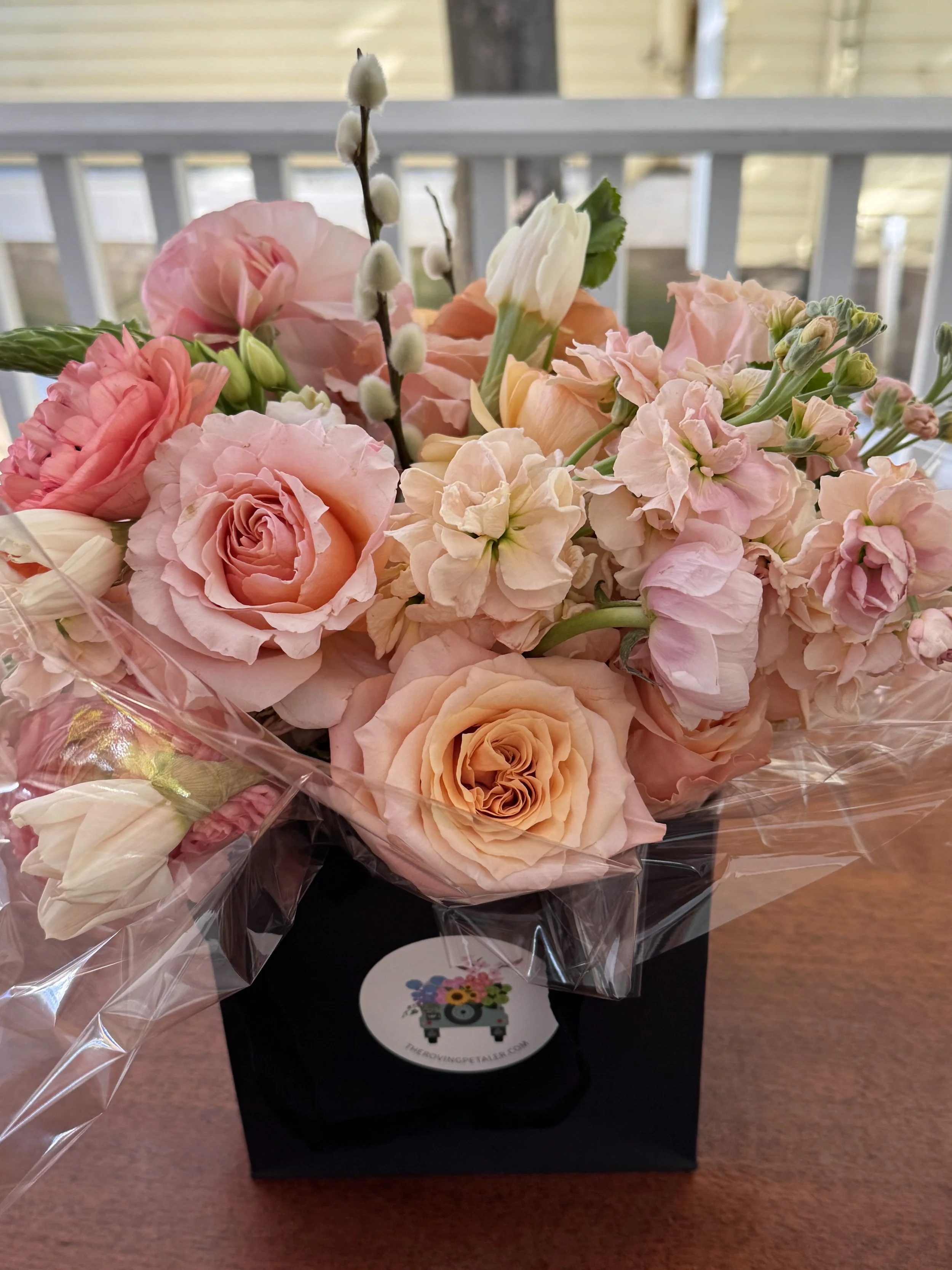 A bouquet of pink, peach, and white roses, and other flowers in a black box with a sticker, wrapped in clear plastic, on a wooden table.