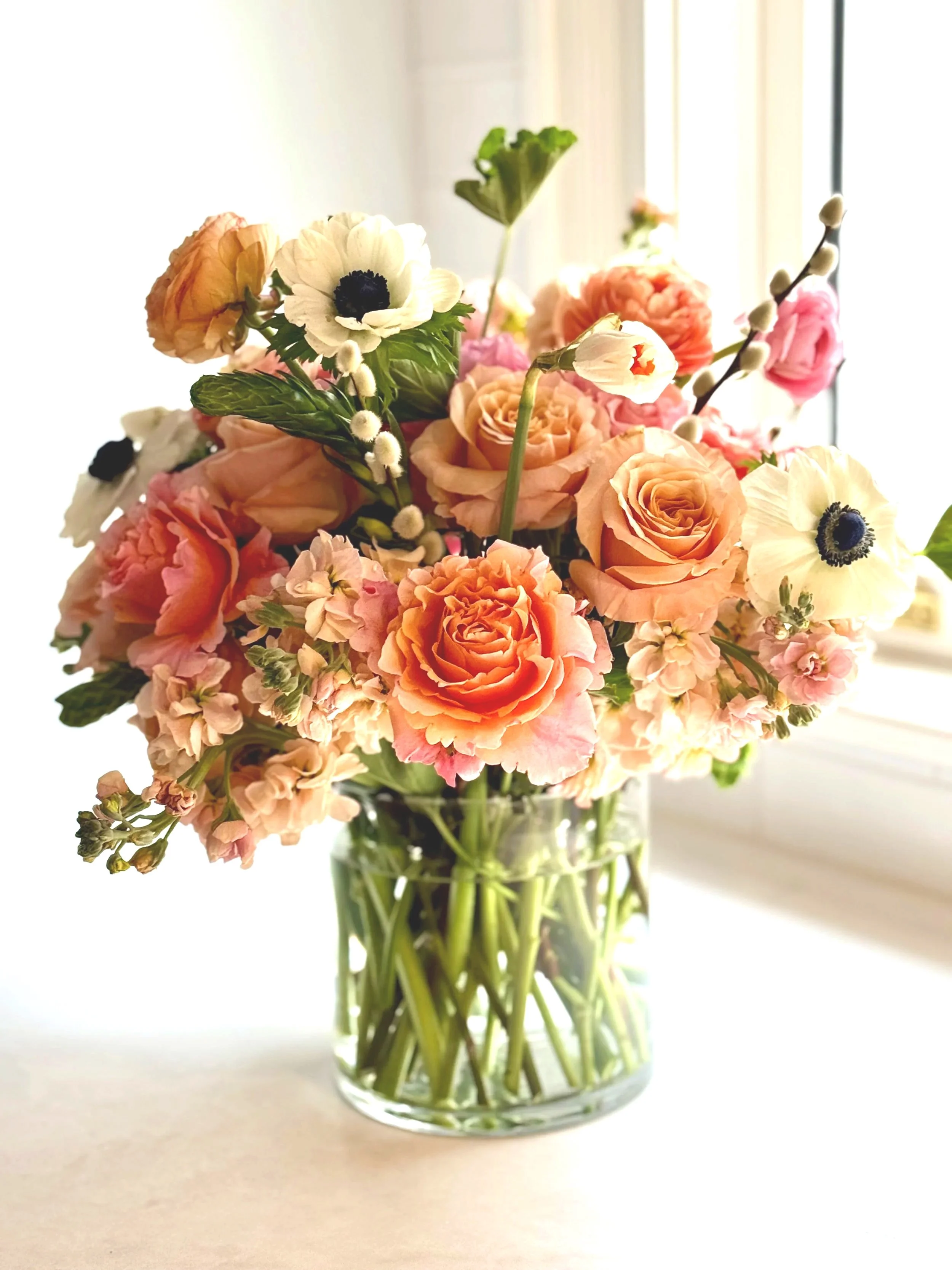 A glass vase with a bouquet of various pink, peach, and white flowers, including roses, anemones, and small blossoms, placed on a white surface near a window with sunlight.