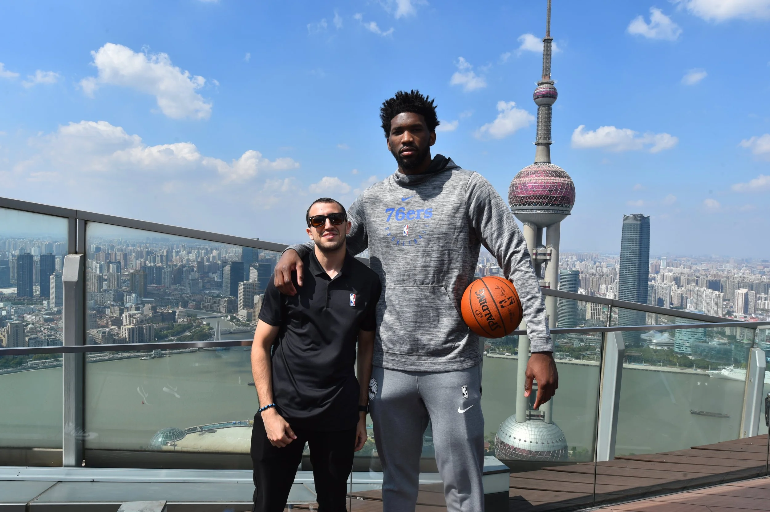 Two men standing on a rooftop balcony with the Shanghai skyline and Oriental Pearl Tower in the background. One man is wearing sunglasses and a black polo shirt with NBA logo, the other is a tall man in a gray NBA hoodie and pants, holding a basketba