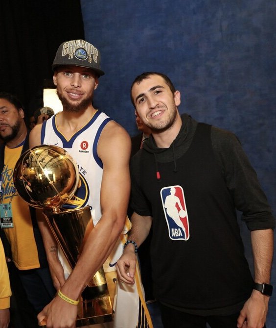 A basketball player wearing a Golden State Warriors championship cap and jersey holding a trophy, standing next to a smiling fan in an NBA hoodie.