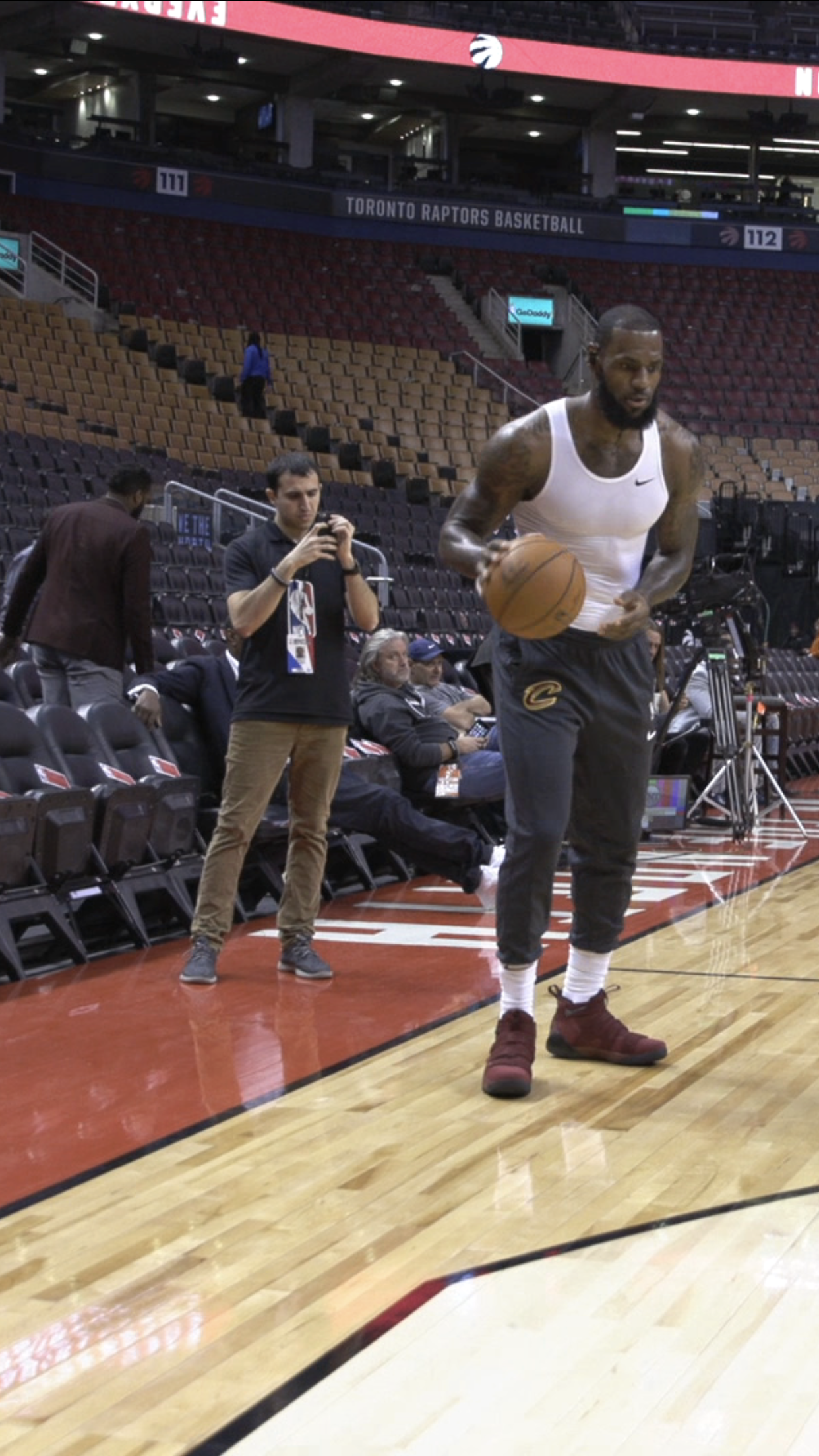 A basketball player dribbling a ball on the court of the Toronto Raptors basketball arena, with people seated and standing nearby, some taking photos or videos.