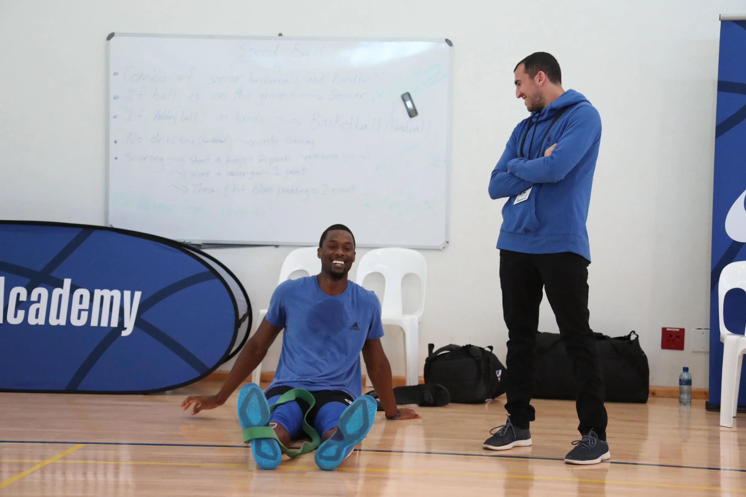 A man sitting on the floor with a smile, wearing a blue athletic shirt, in a sports hall with another man standing beside him, wearing a blue hoodie, white chairs, sports bags, a whiteboard, and a blue banner with the word 'academy' partially visible