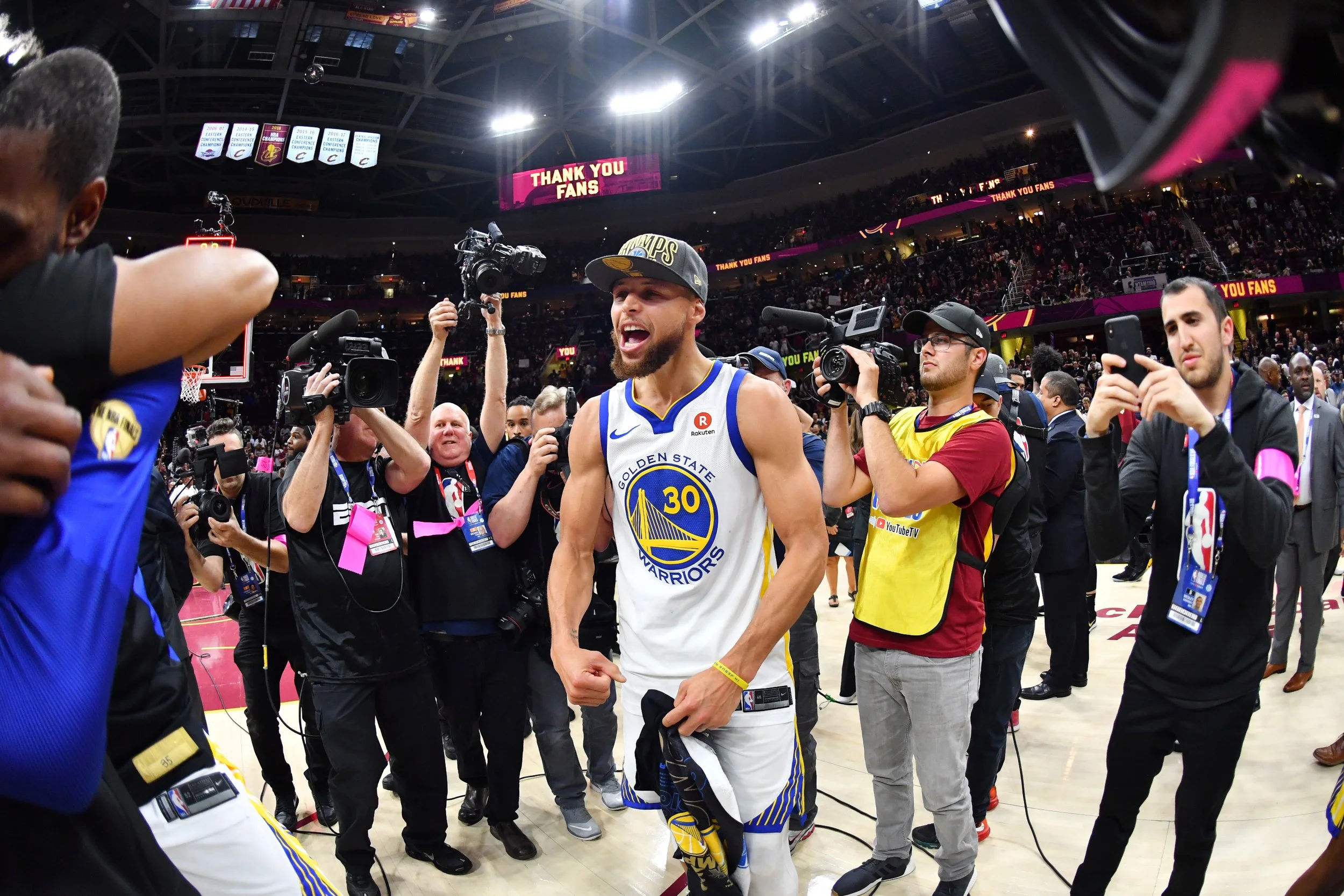 Golden State Warriors player celebrating on the basketball court surrounded by media and photographers, with a scoreboard and banners in the background.