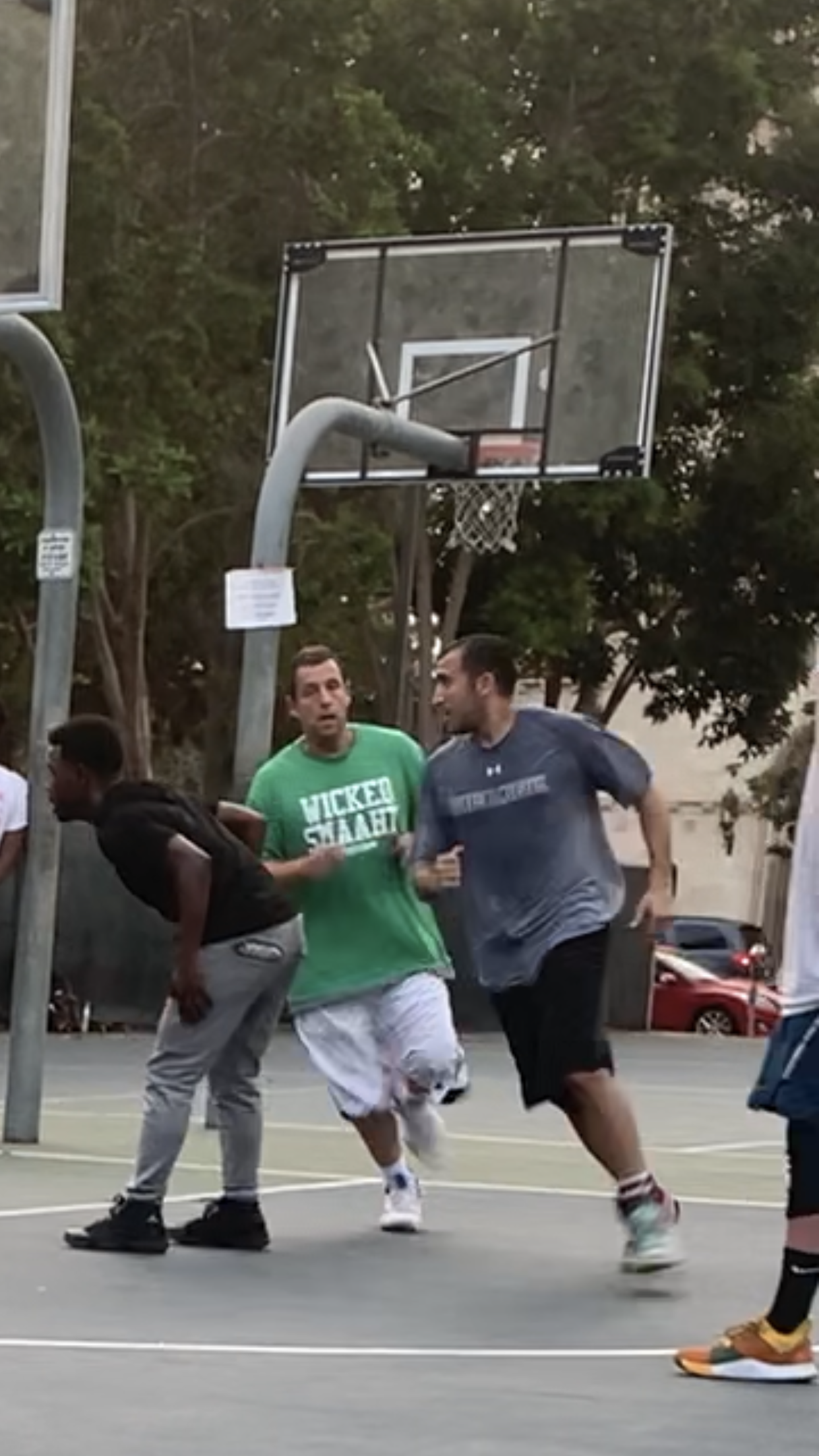 Three men running on an outdoor basketball court with a basketball hoop in the background. One man is wearing a black shirt and gray pants, another is in a green shirt and white shorts, and the third is in a gray shirt and black shorts.