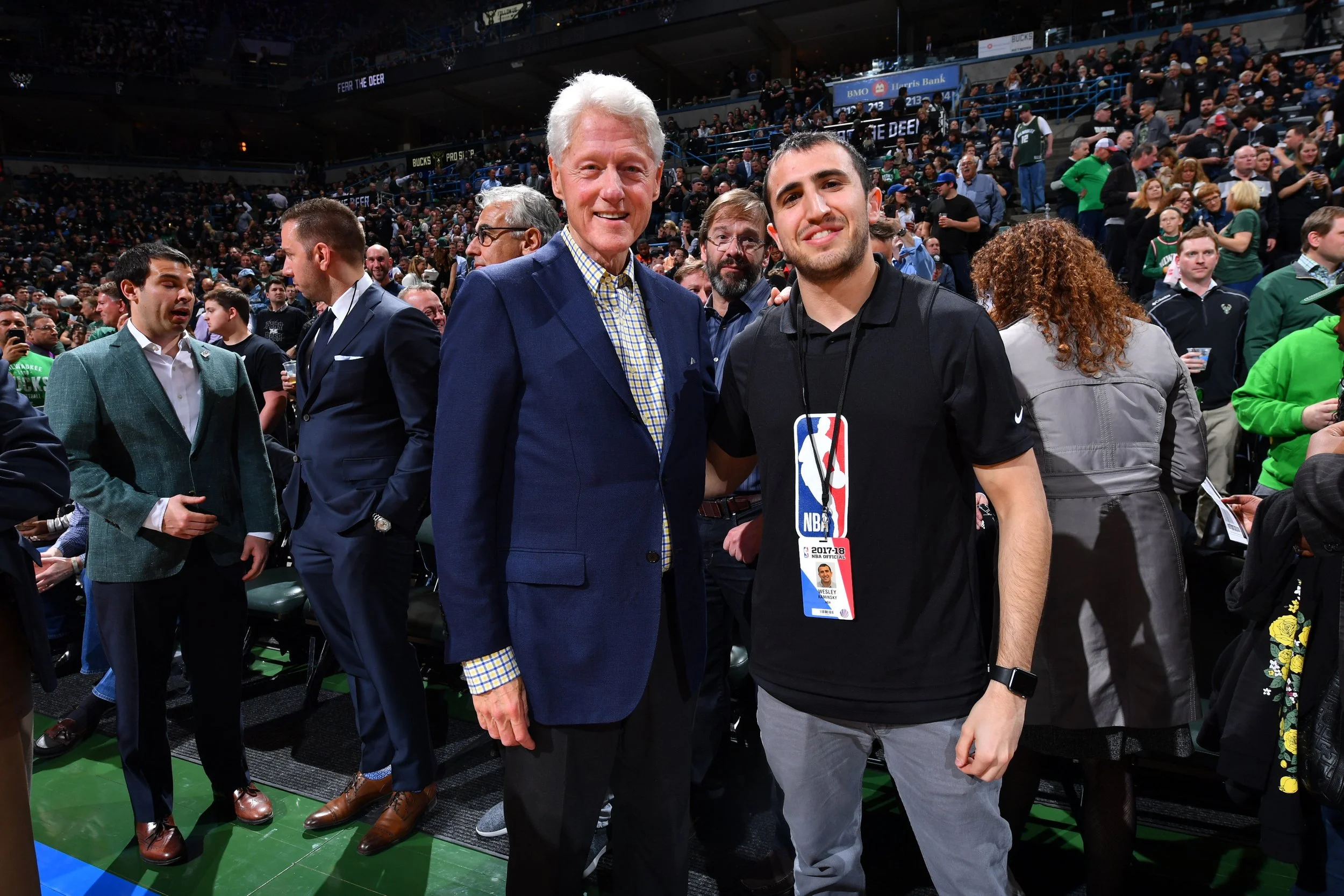 Two men pose for a photo at a crowded NBA event in an indoor sports arena. The man on the left is older with white hair, wearing a blue blazer and checkered shirt. The man on the right is younger with short dark hair, wearing a black NBA T-shirt with