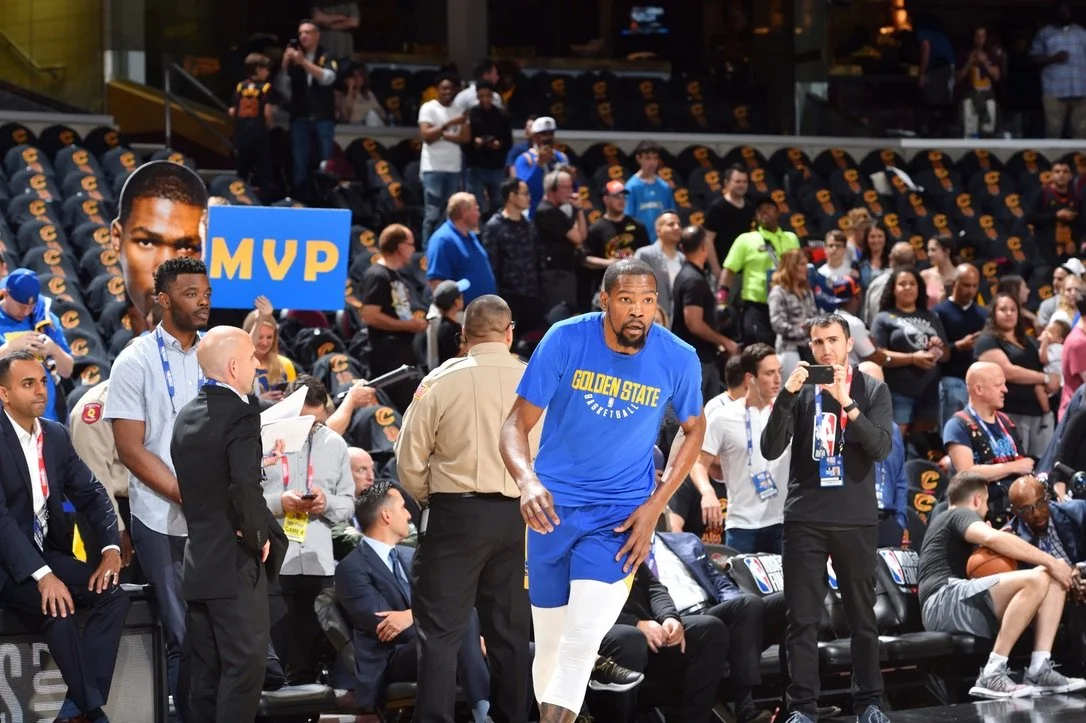 A basketball player in a blue Golden State Warriors uniform walking on a basketball court, with people and photographers in the background during a game or event.