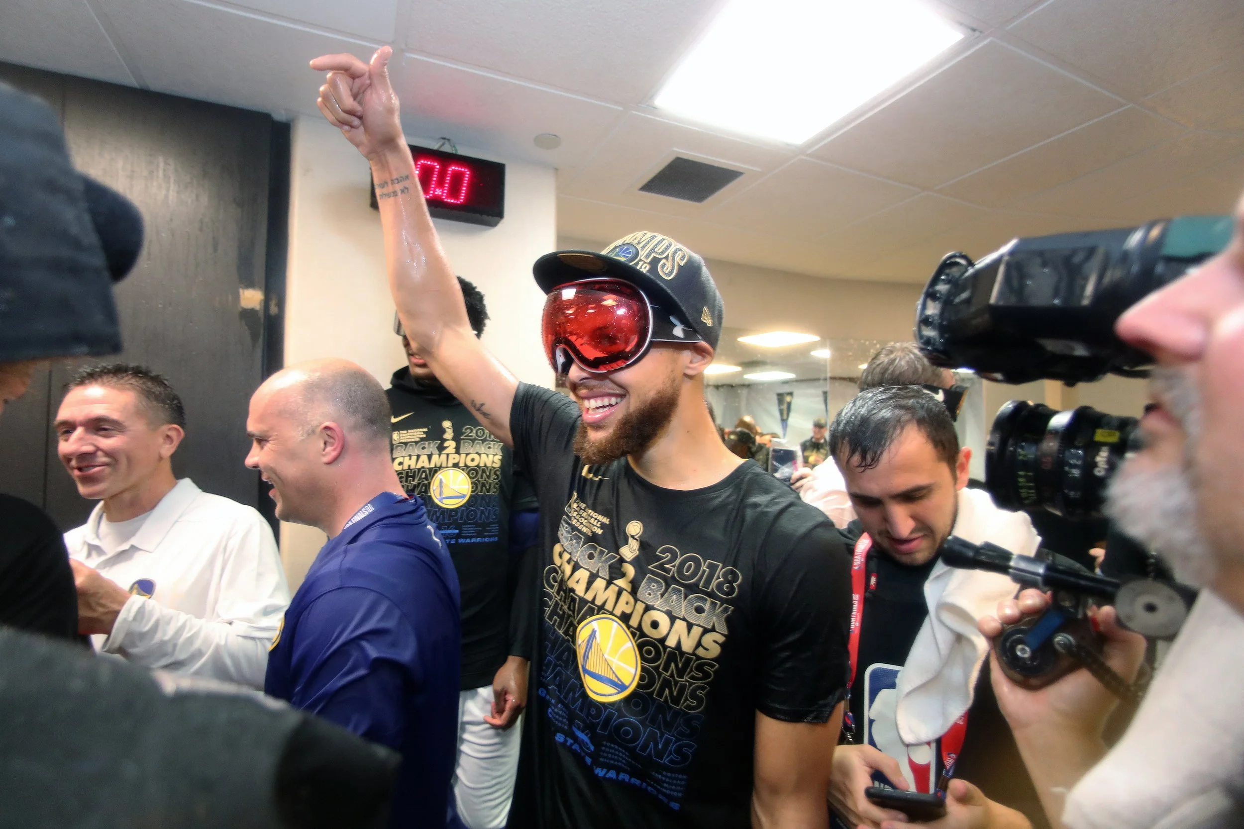 Golden State Warriors player celebrating in a locker room with sunglasses and a hat, surrounded by teammates and media, holding his hand up in victory.