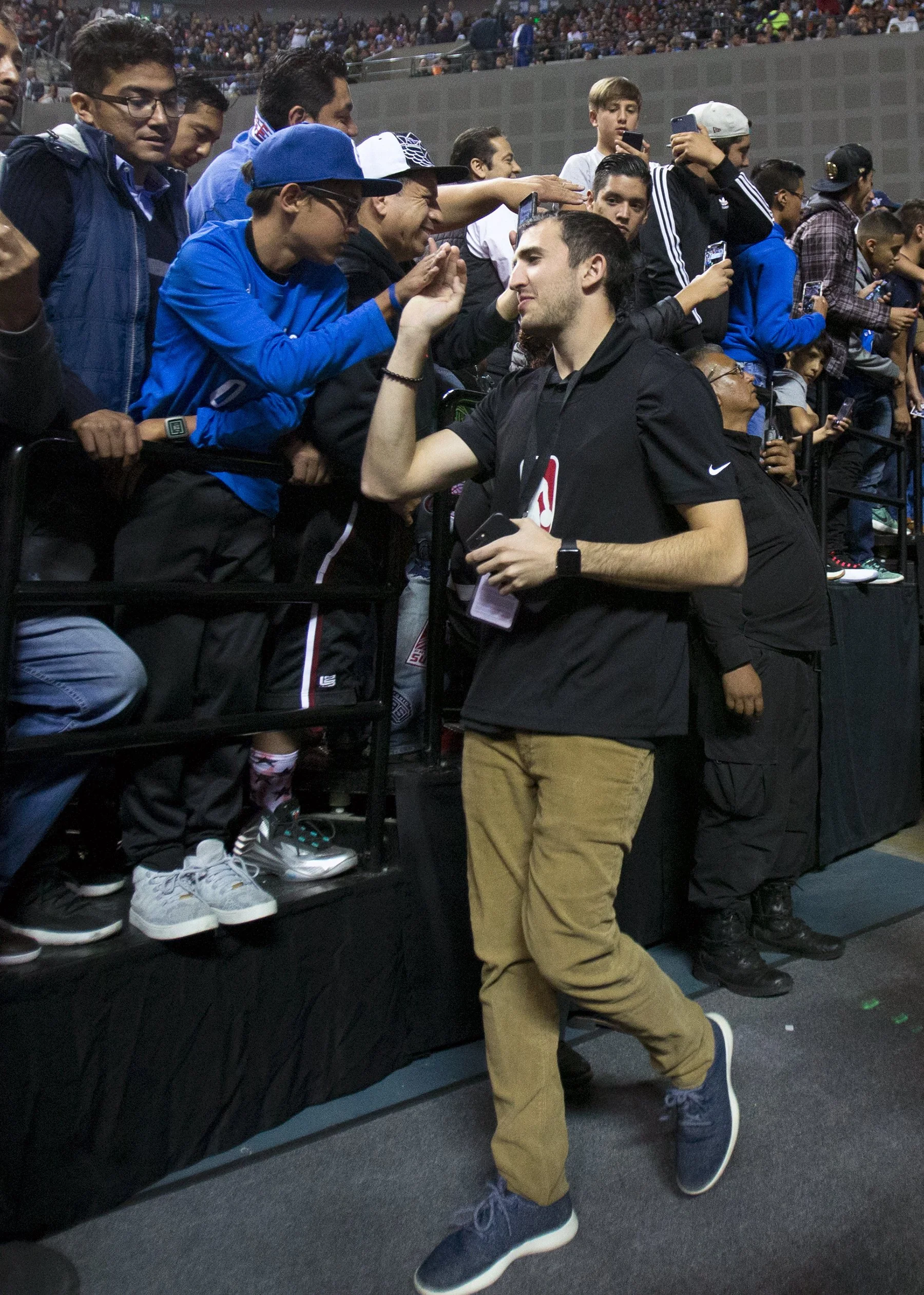 A man in a black hoodie and tan pants gives a high-five to a group of young people standing behind a barrier at a sports arena. The crowd appears excited, and some are taking photos of the moment.