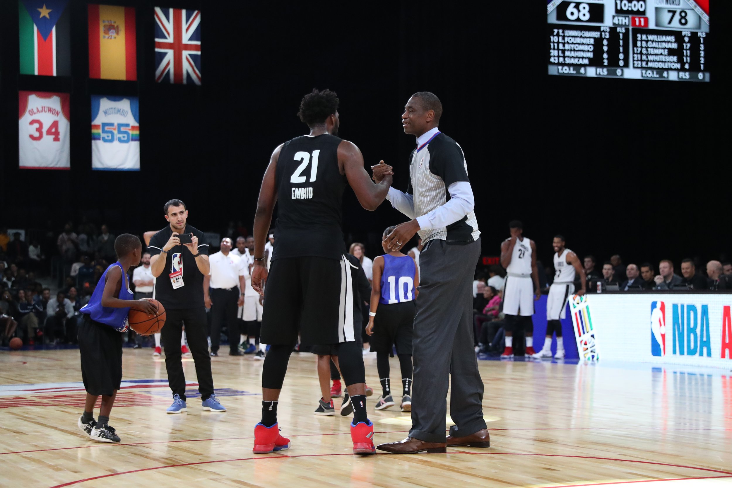 A basketball game scene where a referee is shaking hands with a player wearing a black jersey with the number 21. Other players, a coach, and spectators are visible in the background, with banners and flags hanging above the court.