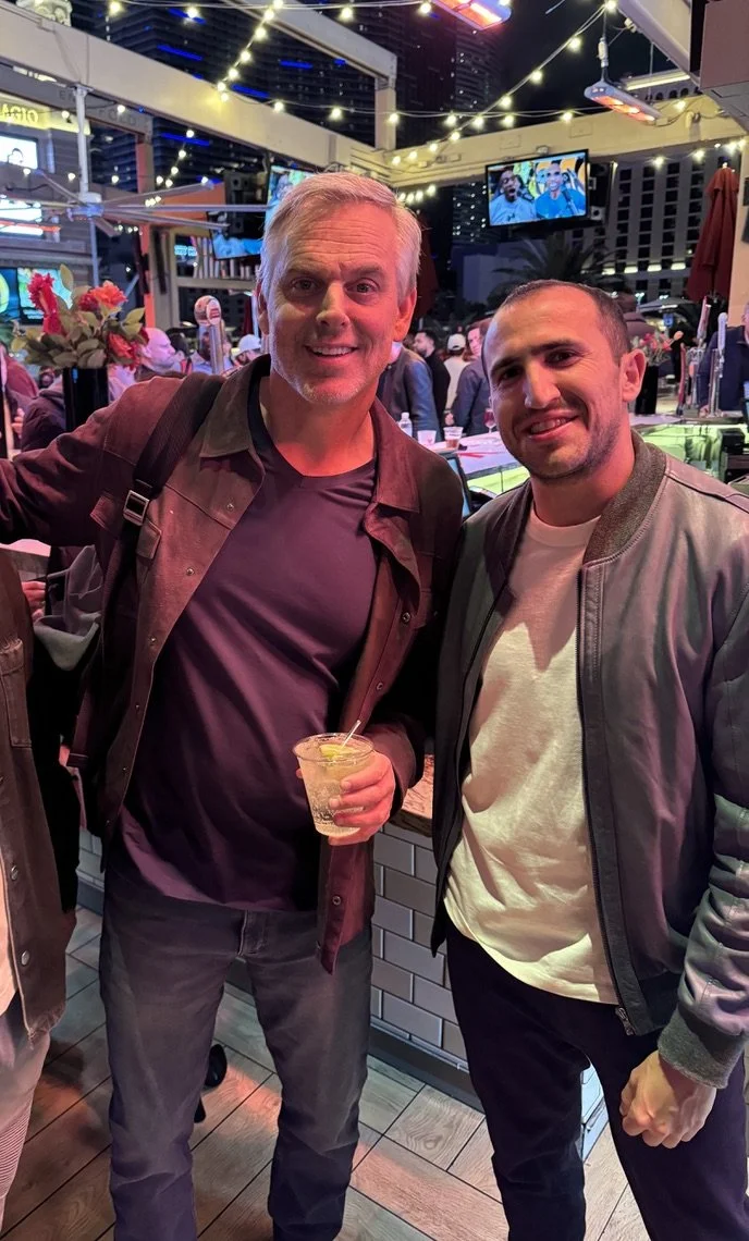 Two men standing together at a lively outdoor bar or restaurant, smiling at the camera. One holds a drink in his hand. Background includes string lights, TV screens, and other people socializing.