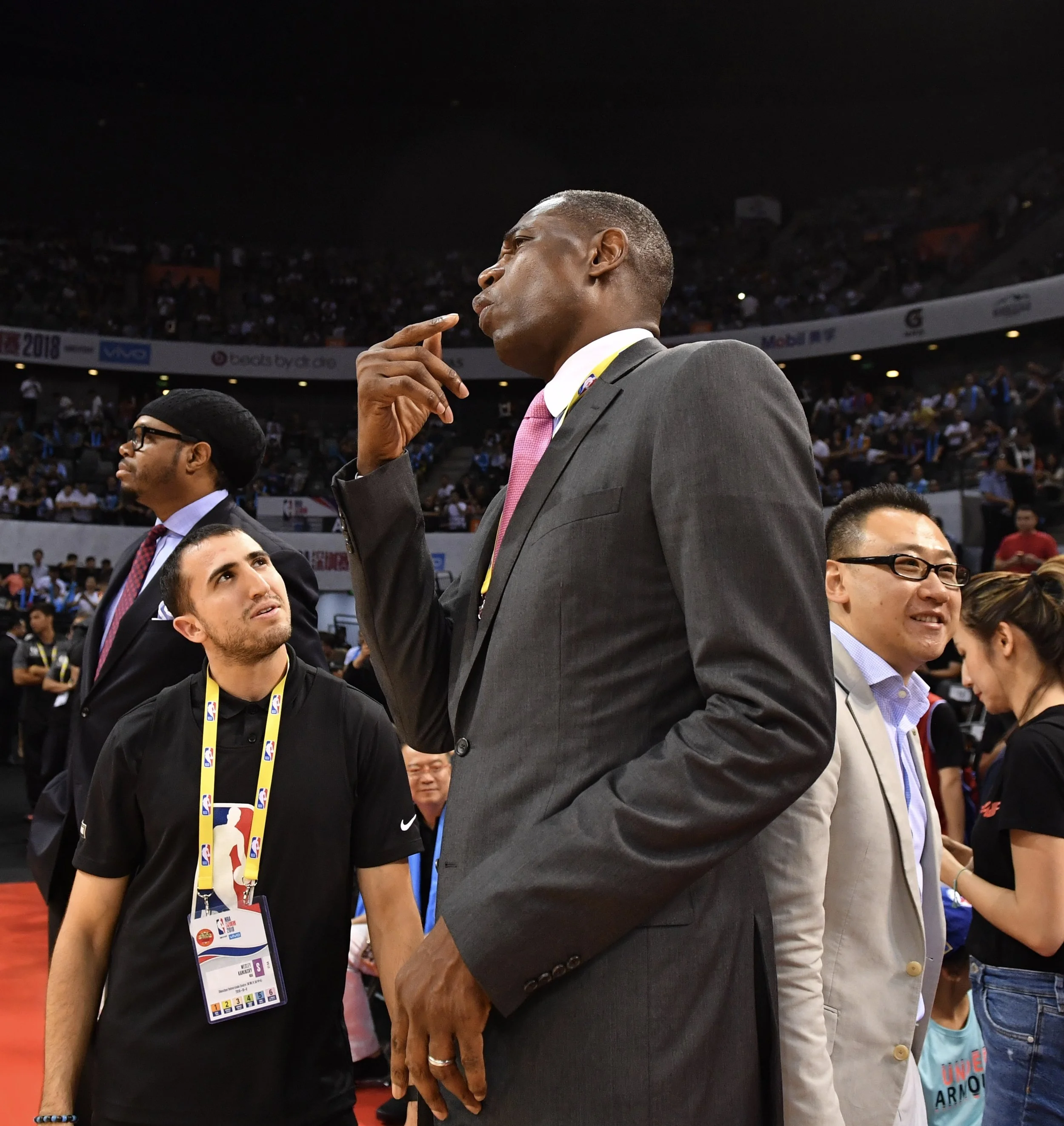 A tall man in a gray suit with pink tie, deep in thought, standing among other people at a sports event in a large indoor arena with many spectators.