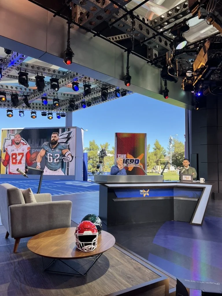 TV studio set for sports talk show with multiple large screens, two hosts, armchairs, sports helmets on a small table, and an outdoor view in the background.