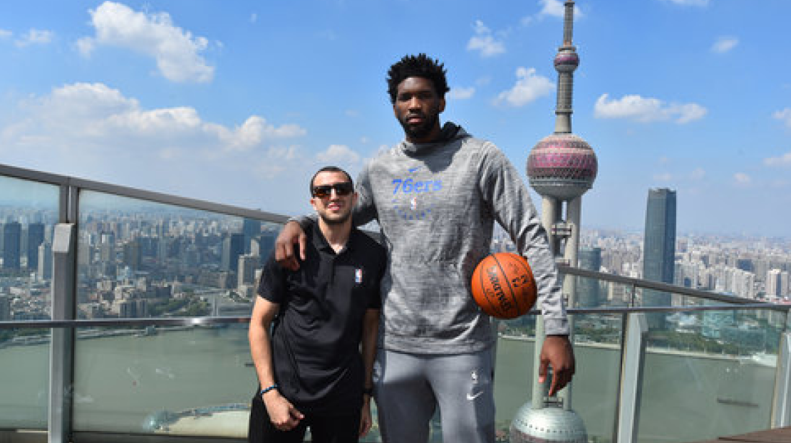 Two men pose on a rooftop with a city skyline in the background, including a tall tower with a spherical design, one man holding a basketball.