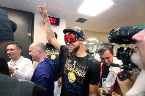 A man wearing a Black Golden State Warriors championship T-shirt, red glasses, and a cap celebrating with his arm raised in a locker room surrounded by smiling teammates and media.