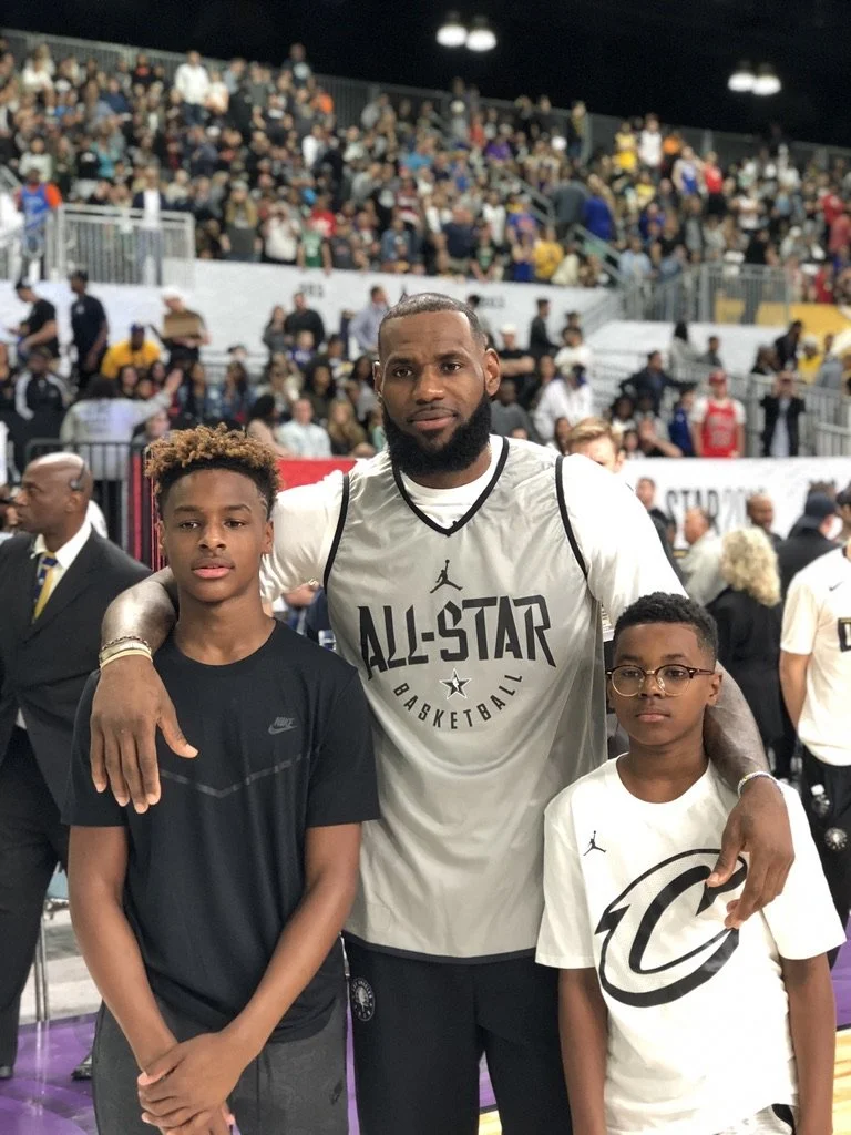 A man with a beard wearing an NBA All-Star basketball jersey stands with two young boys, one wearing a black Nike t-shirt and the other wearing a white t-shirt with a black logo, on a basketball court with a crowd in the stands behind them.