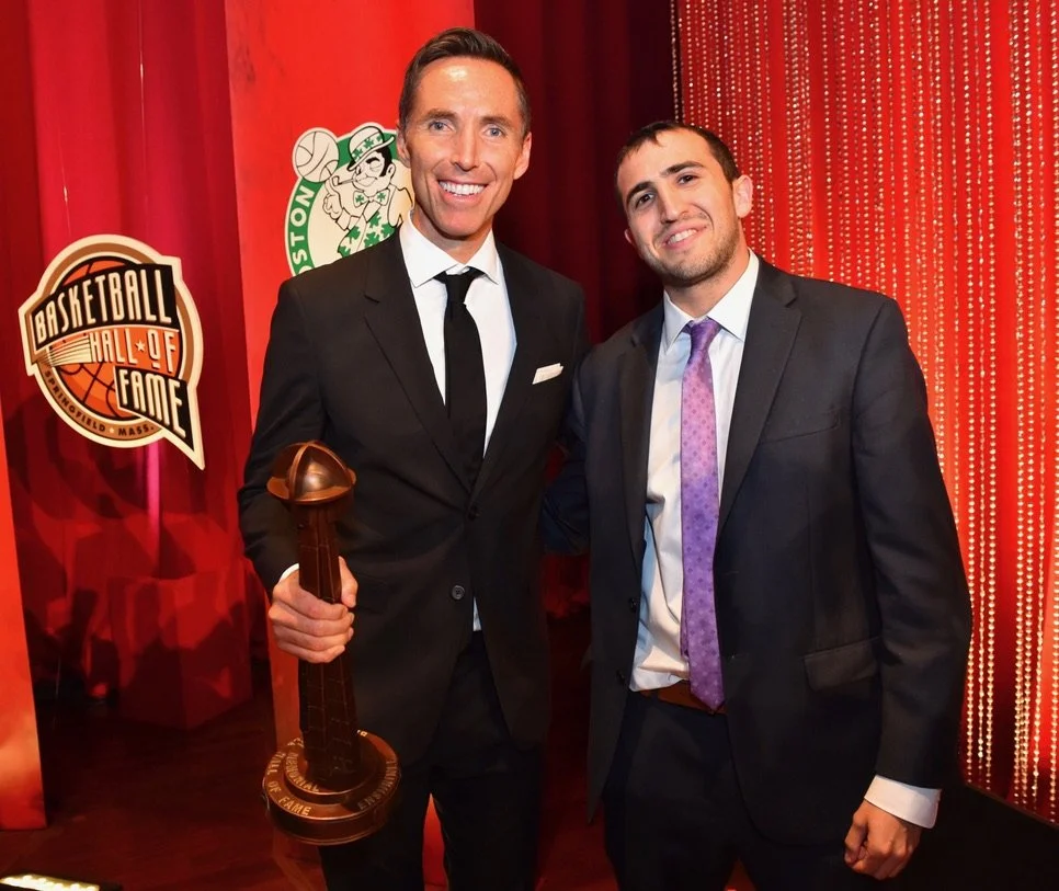 Two men in suits smiling at an awards ceremony with a red curtain backdrop. One man holds a basketball-themed trophy. A sign on the left reads 'Basketball Hall of Fame'.