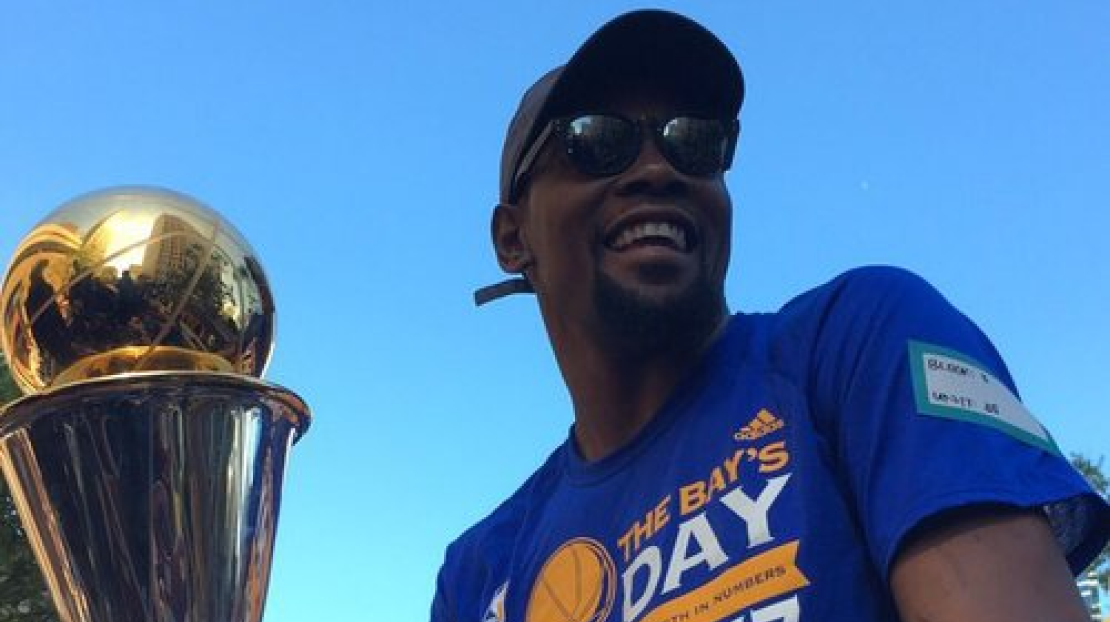 A man in a blue sports shirt with a medal on his arm, wearing sunglasses and a cap, smiling outdoors with a blue sky, holding a trophy with a gold ball on top.