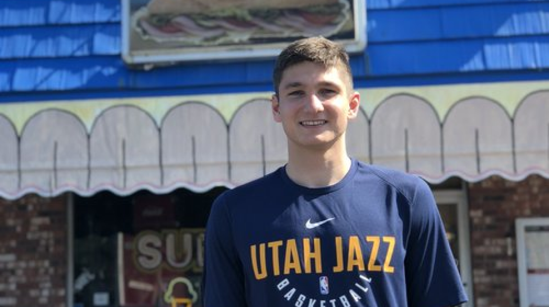Young man smiling outdoors, wearing a navy blue Utah Jazz basketball shirt, standing in front of a building with a colorful awning and a window.