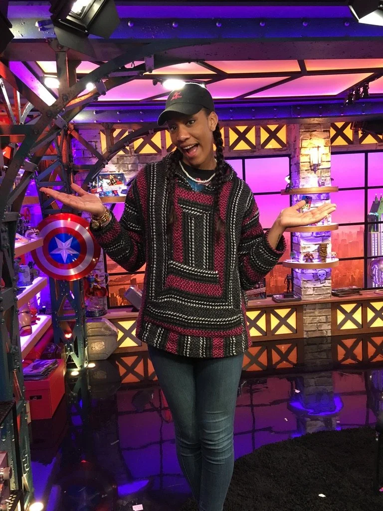 Young woman with braided hair, wearing a black cap, colorful sweater, and jeans, standing in a brightly lit room with shelves and colorful background, holding a Captain America shield in her left hand and gesturing with her right hand.