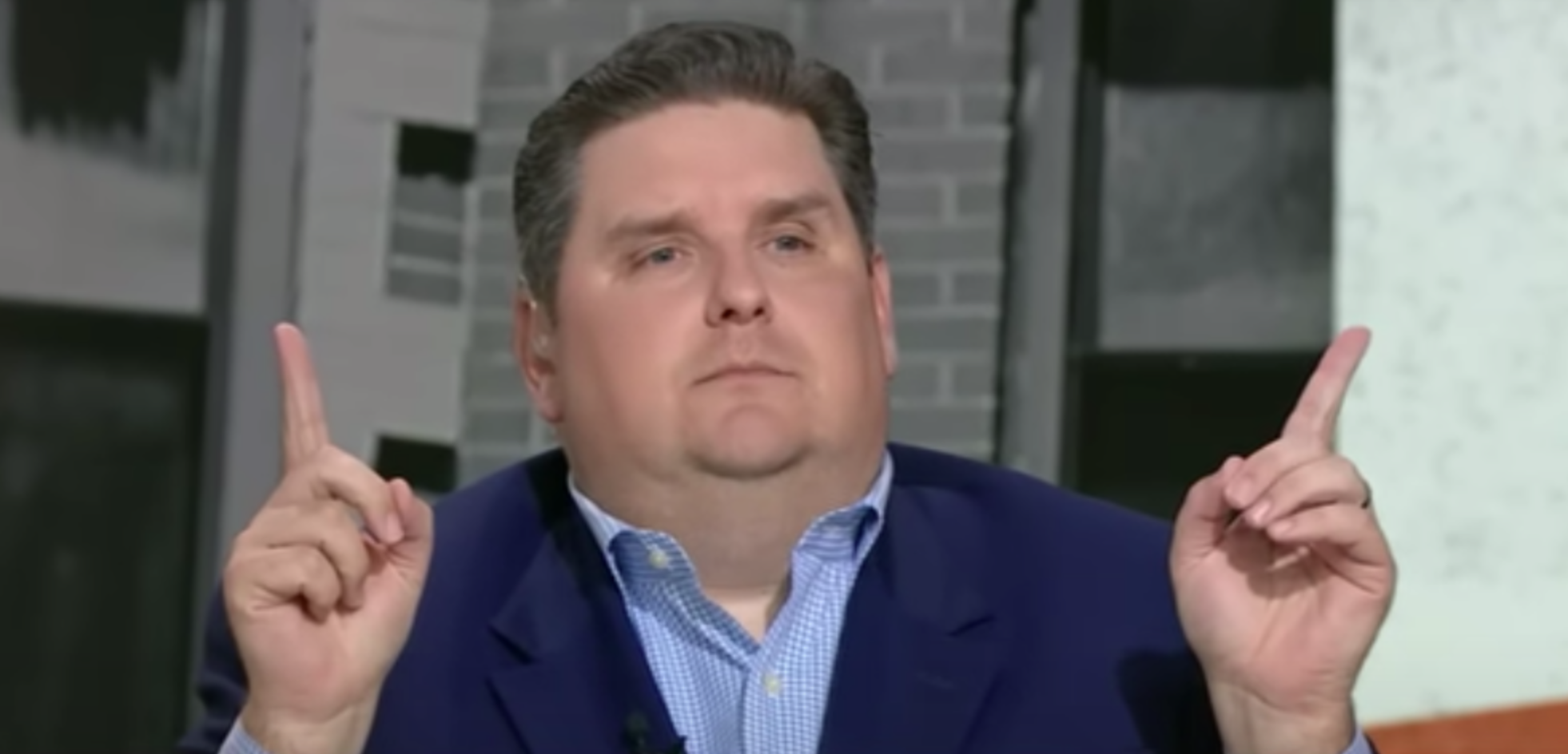 A man with short dark hair, wearing a dark suit jacket and blue collared shirt, is sitting indoors with a neutral expression while raising both hands with index fingers pointing upward.