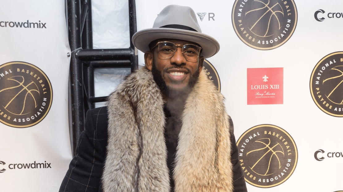 A man with glasses, a fedora, and a fur coat smiling at a sports event with banners of the National Basketball Players Association in the background.