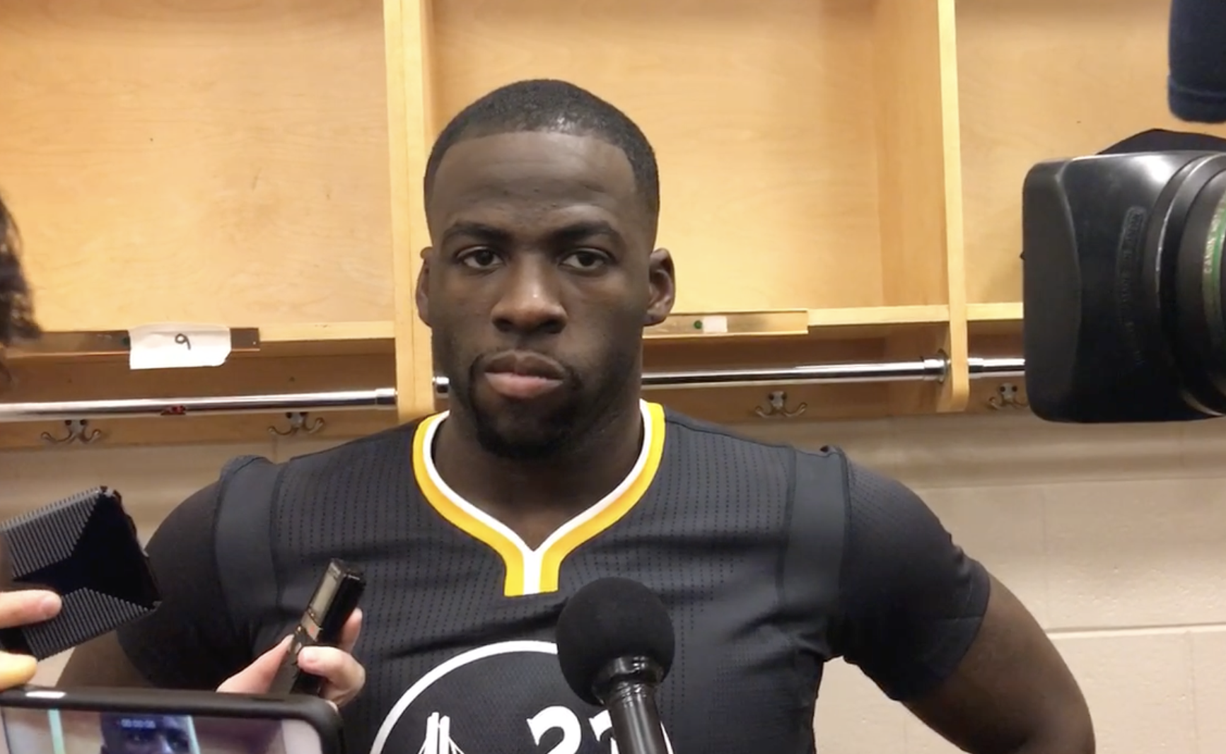 A young African American male athlete with a serious expression, wearing a black sports jersey with yellow and white accents, speaking into a microphone in a locker room with wooden lockers in the background.