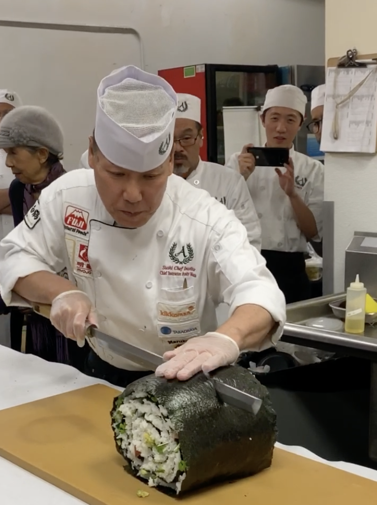 Chef slicing large sushi roll wrapped in seaweed with onlookers in a kitchen setting