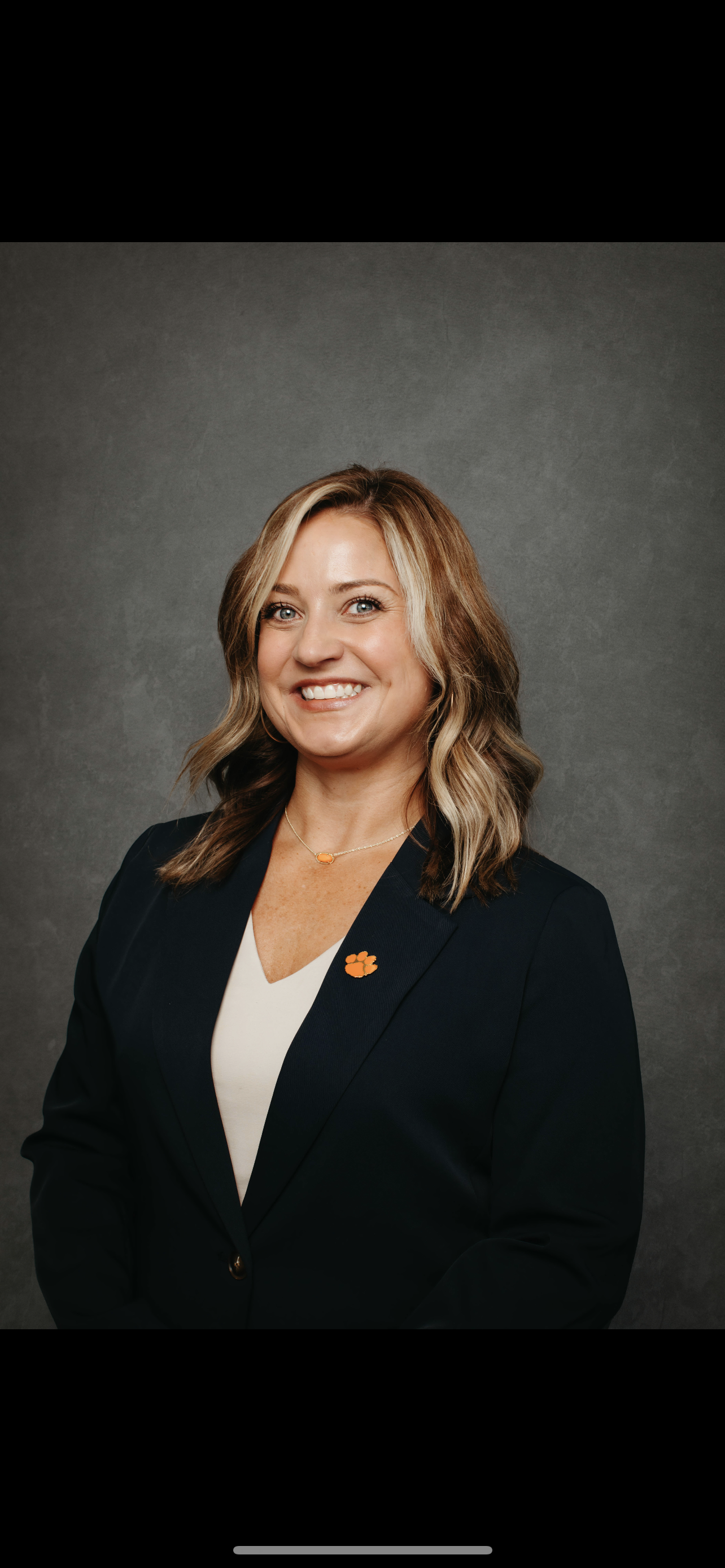 A woman with blonde hair wearing a dark blazer with an orange paw print pin, a necklace, and a cream top, smiling against a gray background.