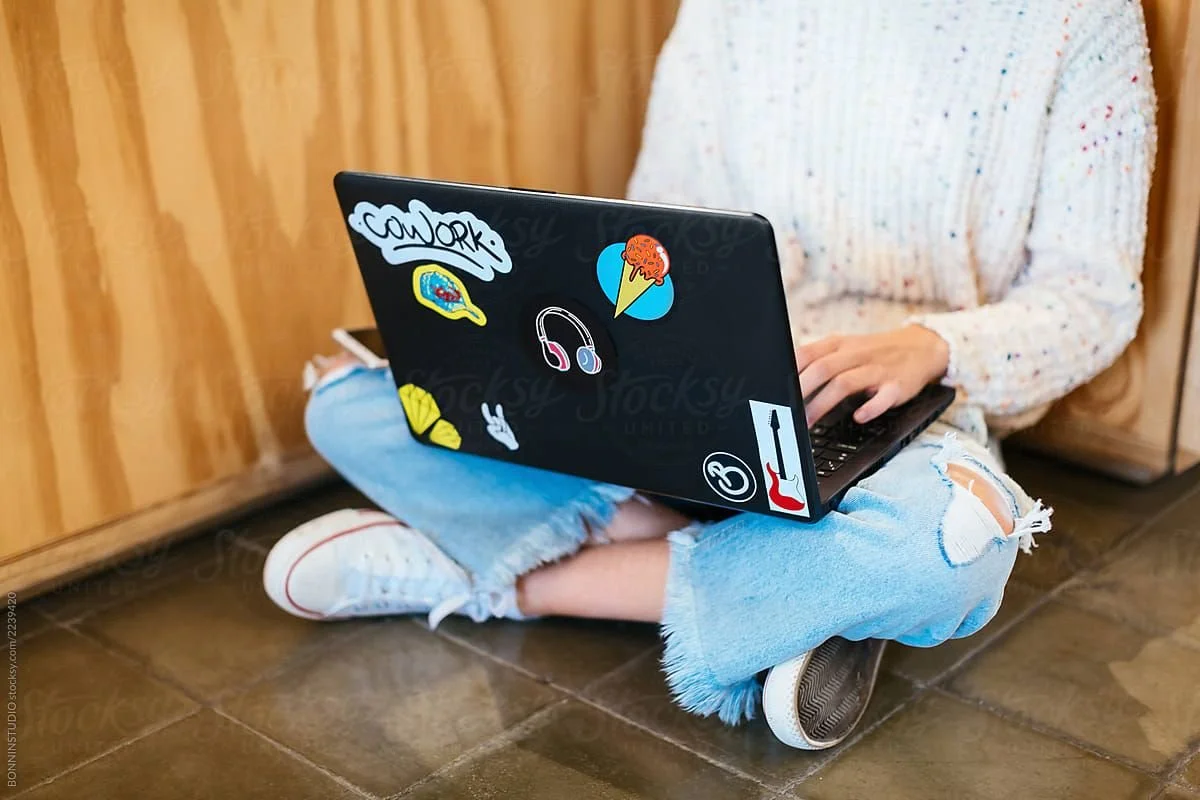 Person sitting cross-legged on the floor using a laptop decorated with stickers, wearing ripped jeans, a cozy sweater, and sneakers.
