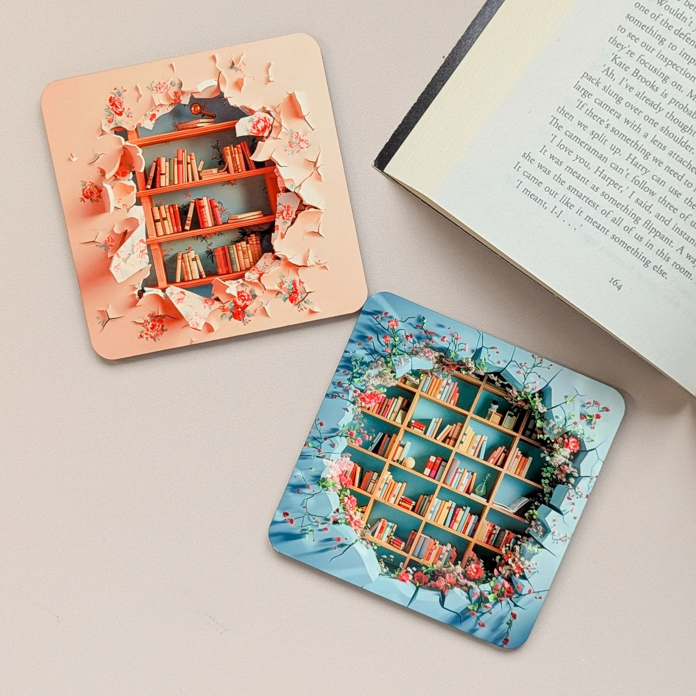 Two decorative coasters featuring bookshelves, one pink with a hole appearing as if torn through, and one blue with a similar design, placed on a light-colored surface next to an open book.