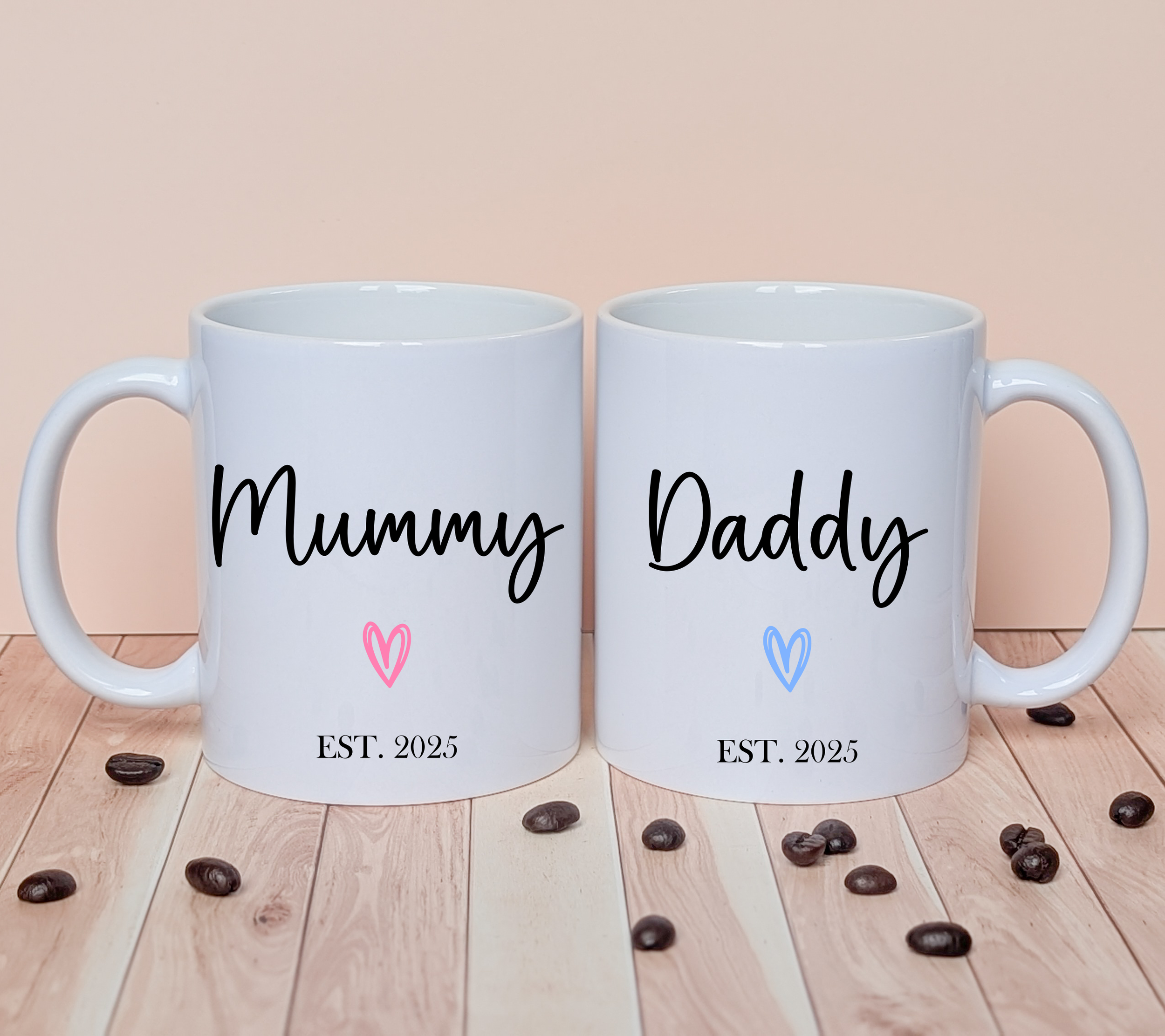 White ceramic mugs with personalised text reading Mummy and Daddy, each featuring a pink or blue heart and ‘Est. 2025’, displayed on wooden surface