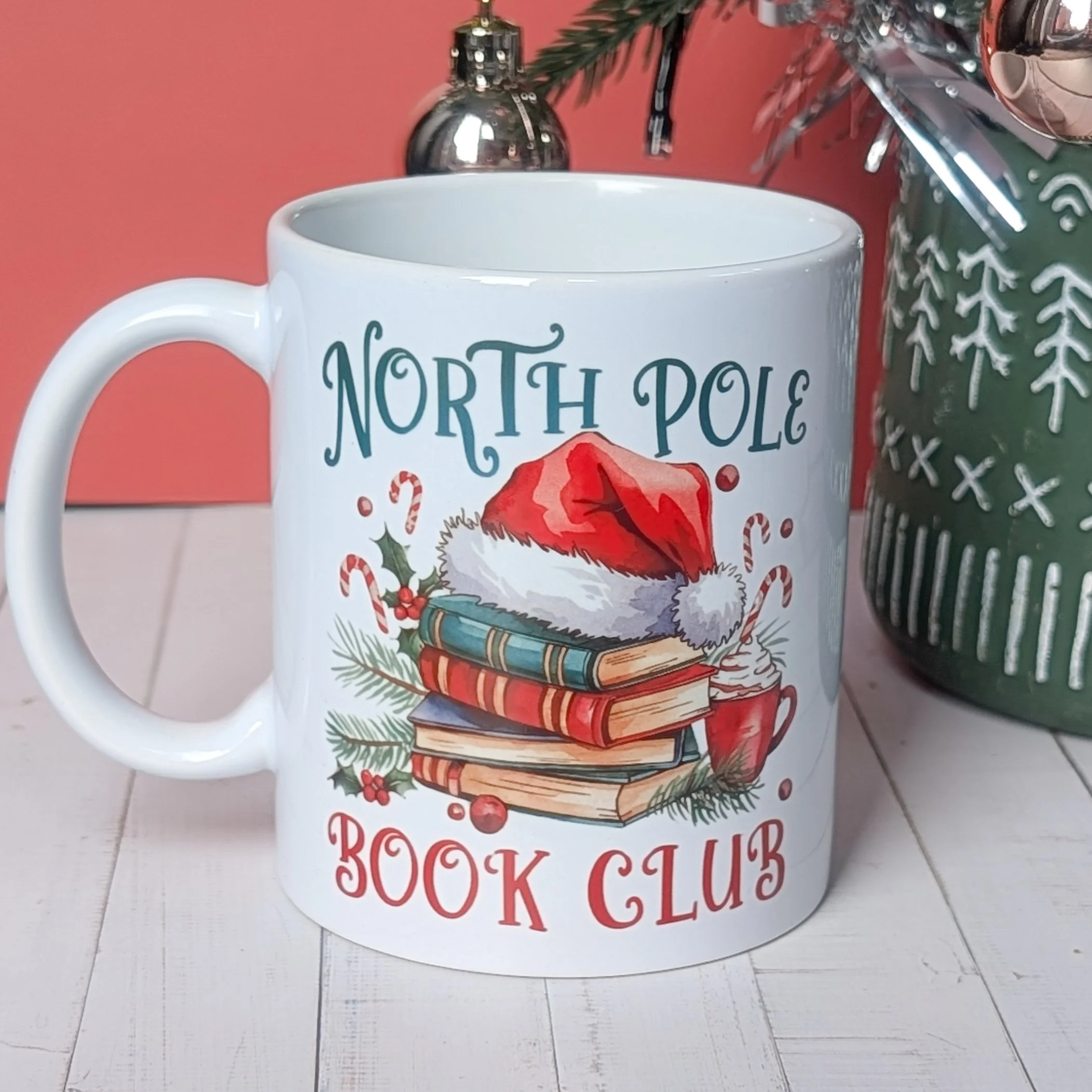 White ceramic mug with ‘North Pole Book Club’ design featuring a Santa hat, festive books, candy canes and cocoa on a wooden background.