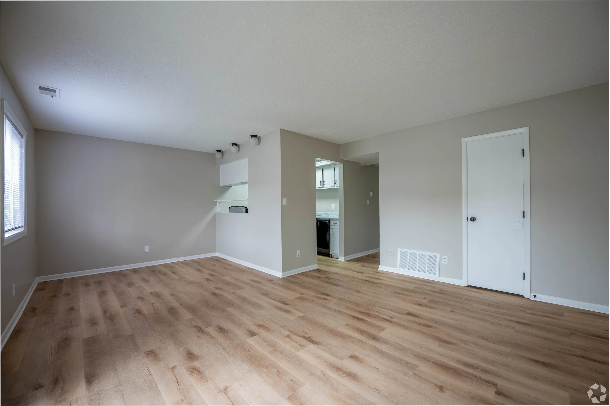 Empty living room with hardwood floors, beige walls, a window with blinds, and an open doorway leading to a kitchen.