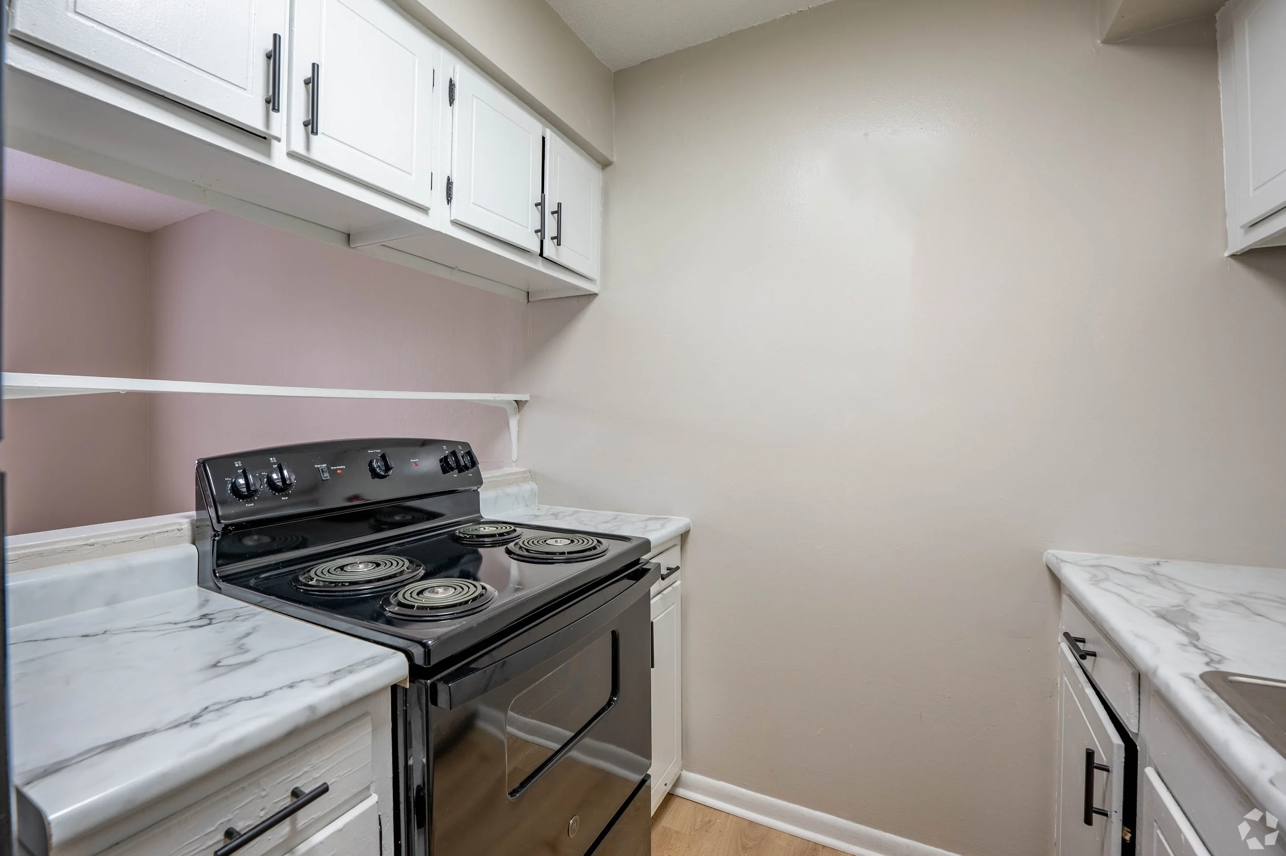 An empty kitchen with white cabinets, marble countertops, and a black electric stove with four burners.