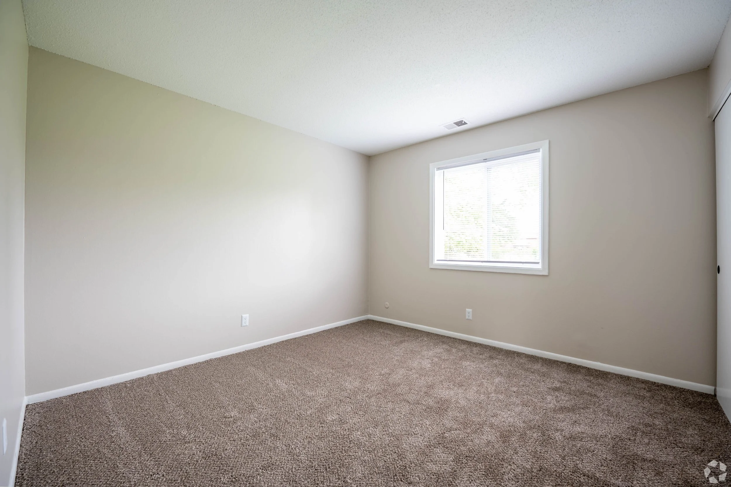 Empty bedroom with beige walls, brown carpet, a window with blinds, and a sliding closet door.