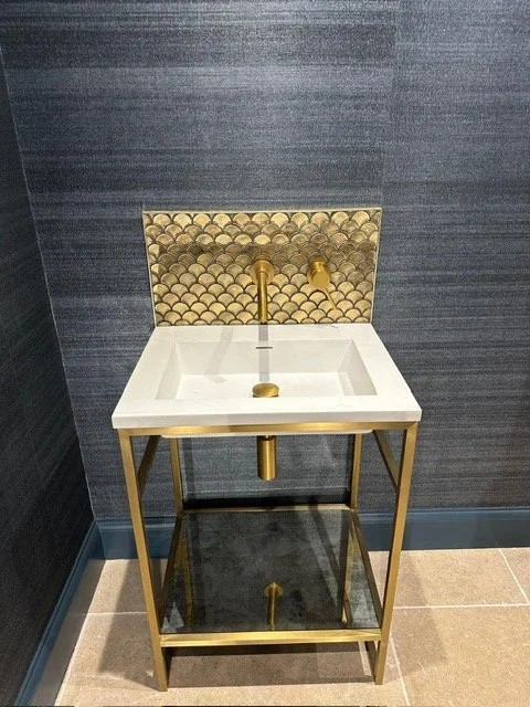 A small white and gold sink with a black shelf underneath, positioned against a dark textured wall with a decorative gold-tiled backsplash.
