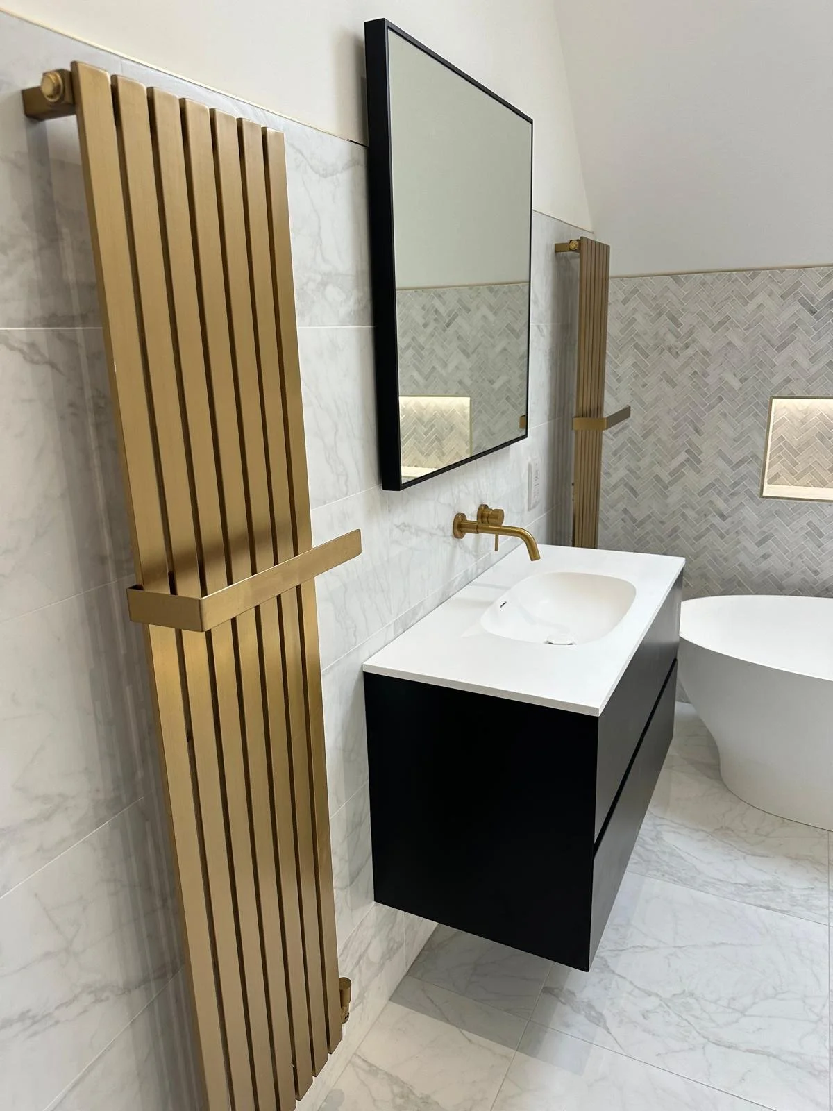 Modern bathroom with black floating vanity, gold fixtures, large mirror, vertical gold radiator, and freestanding bathtub, marble tiling.