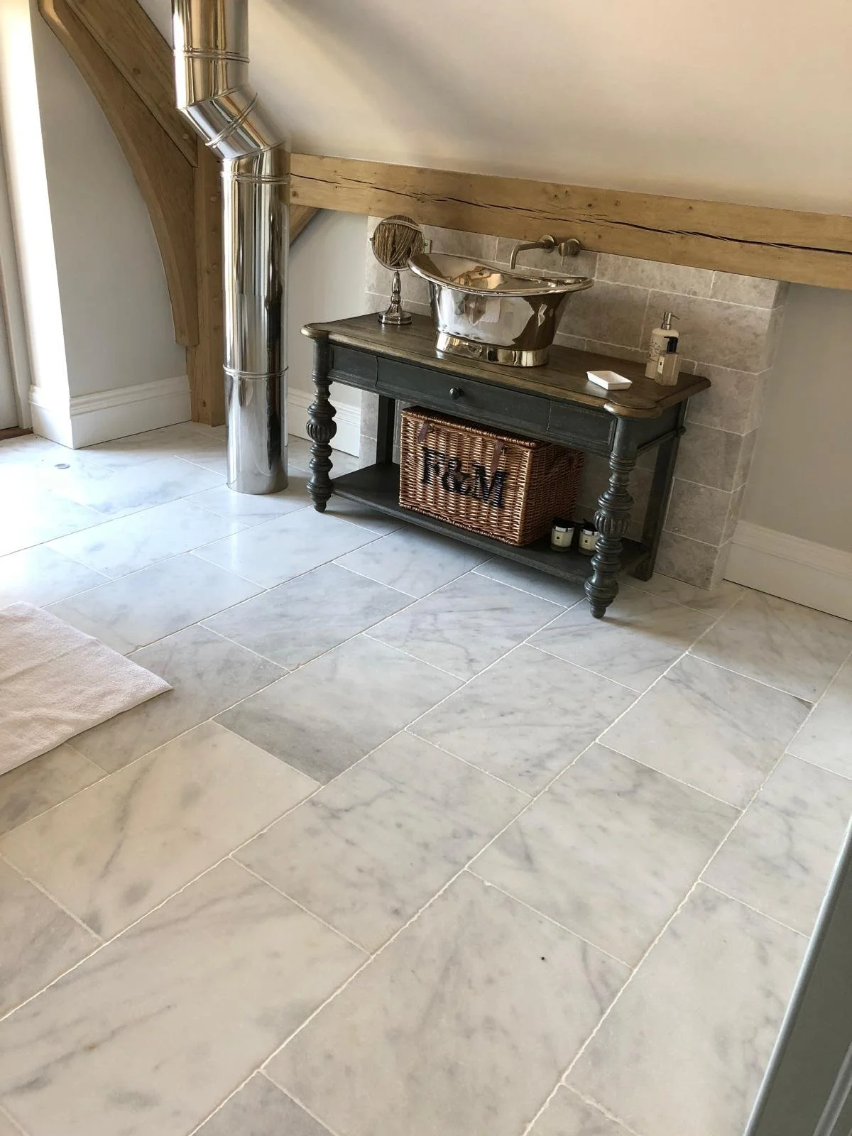 Small black table with a woven basket and three small candles underneath, a shiny metallic bath or basin on top, a small mirror, a soap dispenser, and a lamp, set against a tiled wall, with marble tiles on the floor and visible wooden beams and a met