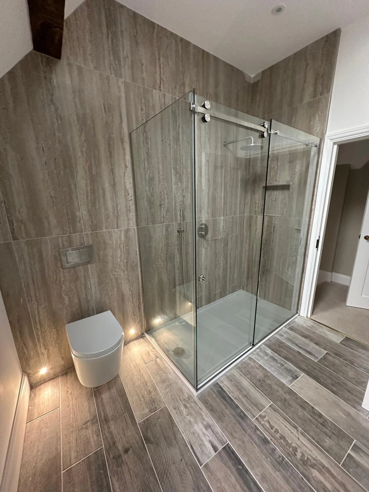 Modern bathroom with a walk-in shower enclosed by glass doors, gray tile walls and floor, a wall-mounted toilet, and subtle lighting.