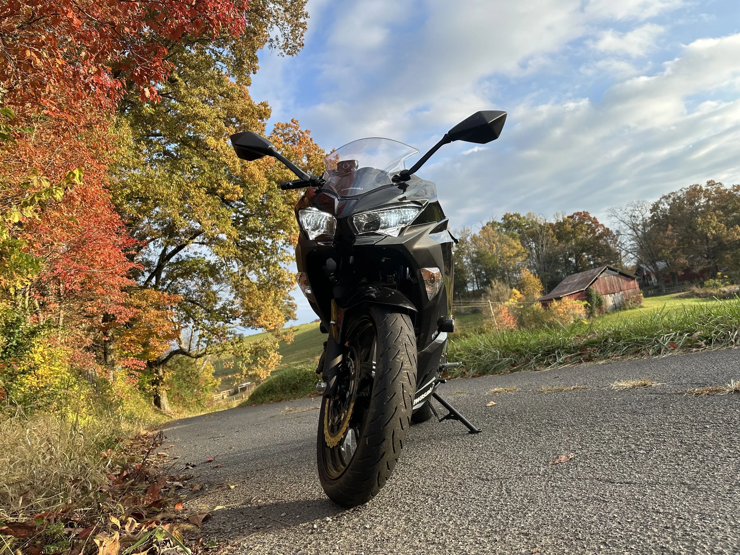A black motorcycle parked on the side of a road surrounded by autumn trees with red, orange, green, and yellow leaves, under a partly cloudy sky.