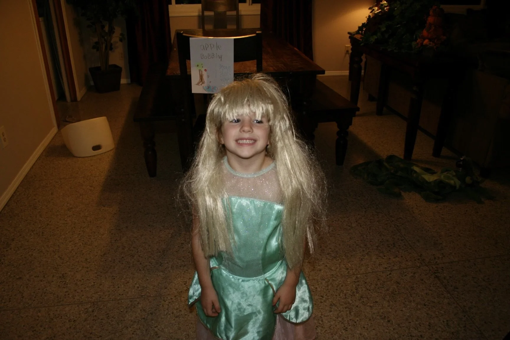 A young girl dressed as Rapunzel in a green princess costume with a long blonde wig, smiling and standing in a living room.