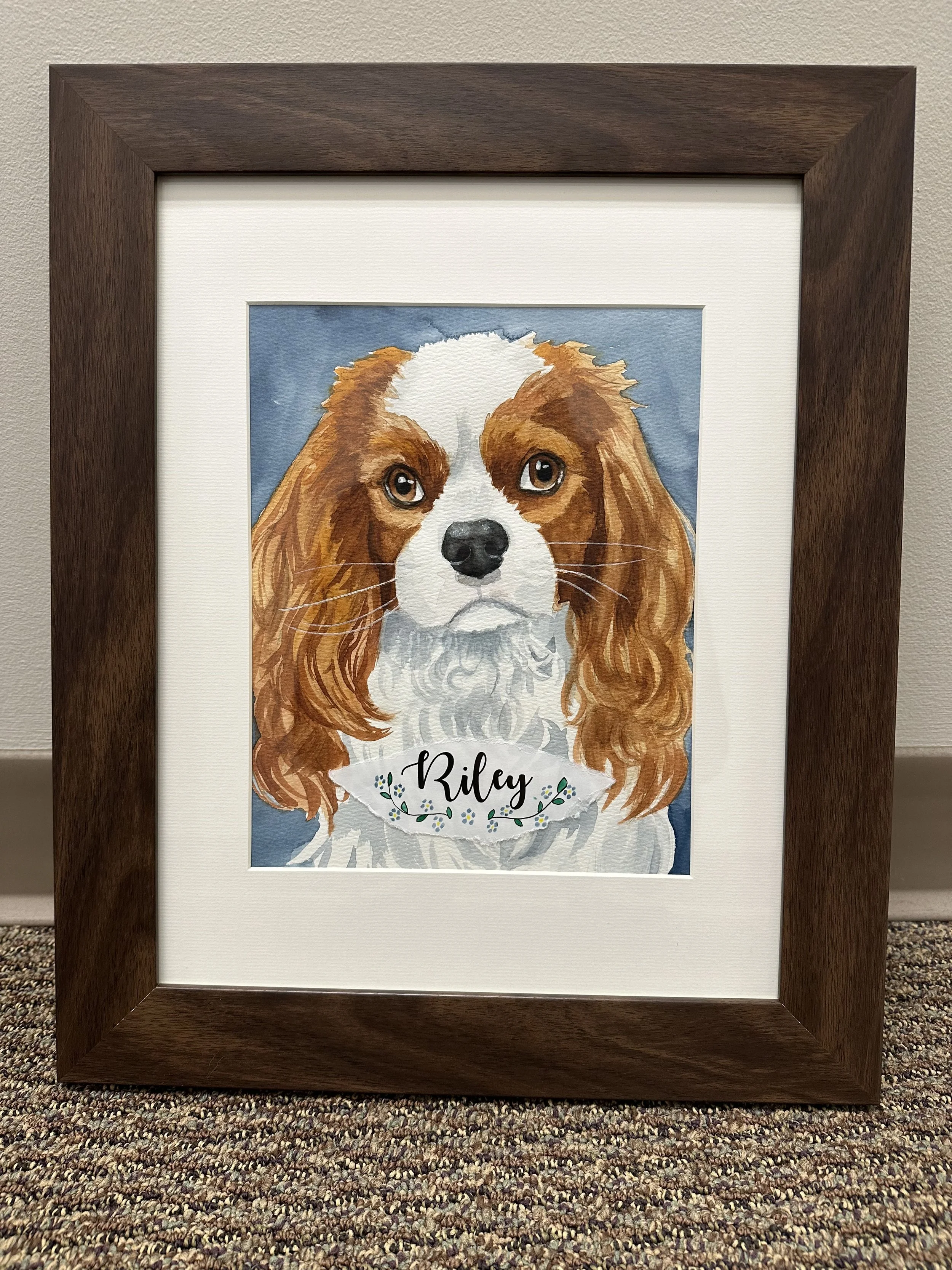 Watercolor painting of a dog with brown and white fur, long ears, and big brown eyes, framed in a dark wooden frame on a textured carpet.