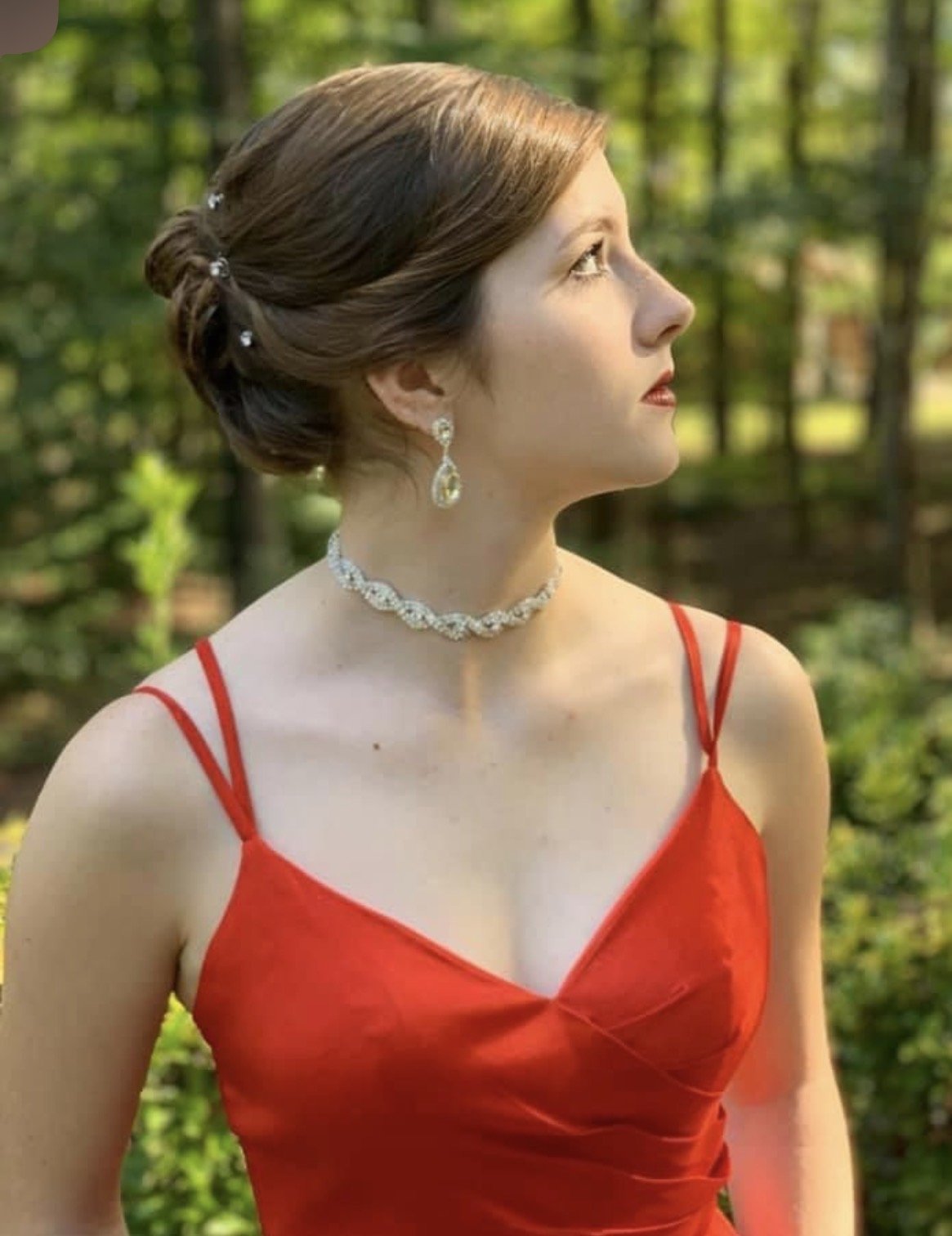 A woman in a red dress with spaghetti straps, wearing diamond jewelry and an elegant updo hairstyle, standing outdoors in a wooded area.