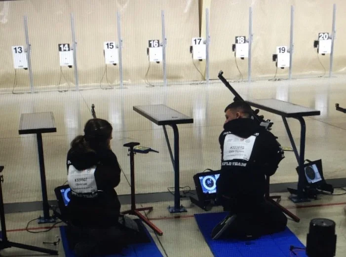 Two people kneeling on blue mats in front of computer monitors at indoor shooting range, with targets numbered 13-20 in the background.
