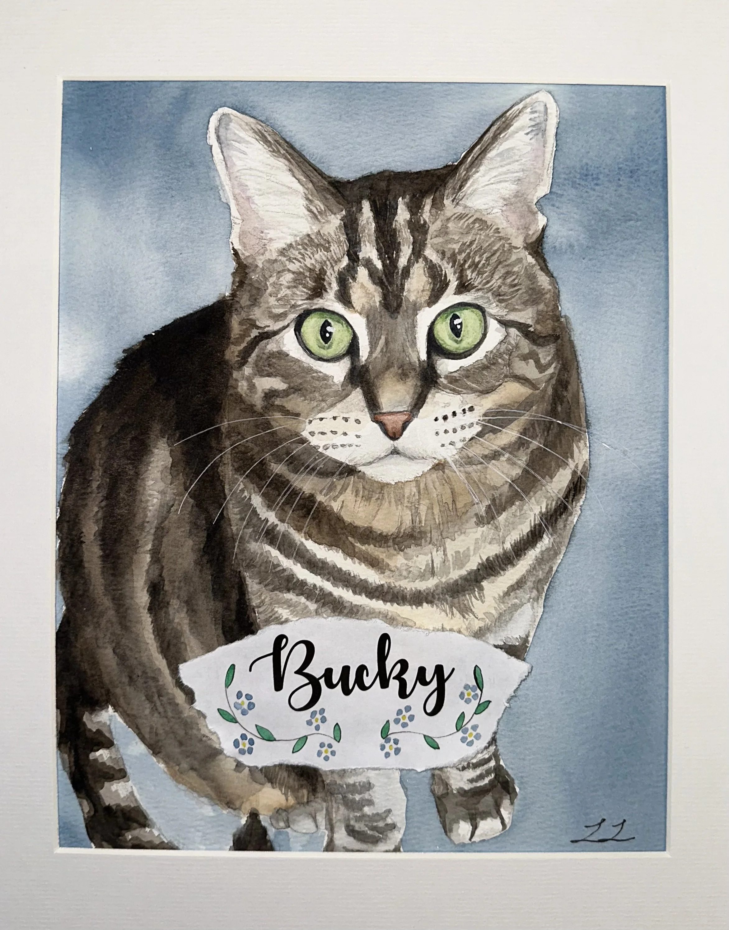 Watercolor painting of a tabby cat with green eyes. The cat is looking at the viewer with a white, black, and brown fur pattern. There is text below the cat that reads 'Bucky' with decorative elements surrounding the name.