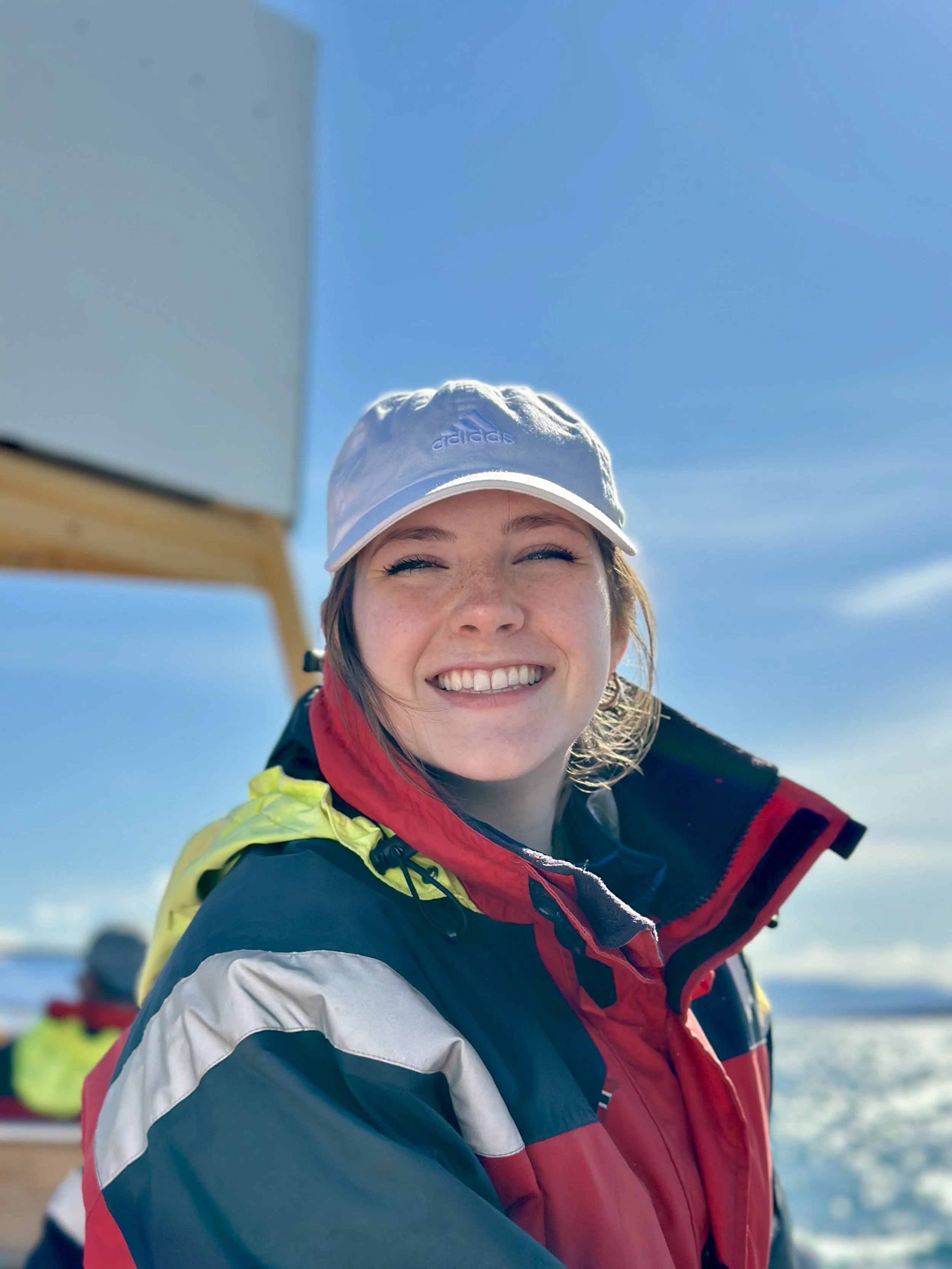 Close-up of a smiling woman wearing a white Adidas cap and a colorful winter jacket, outdoors under a clear blue sky.